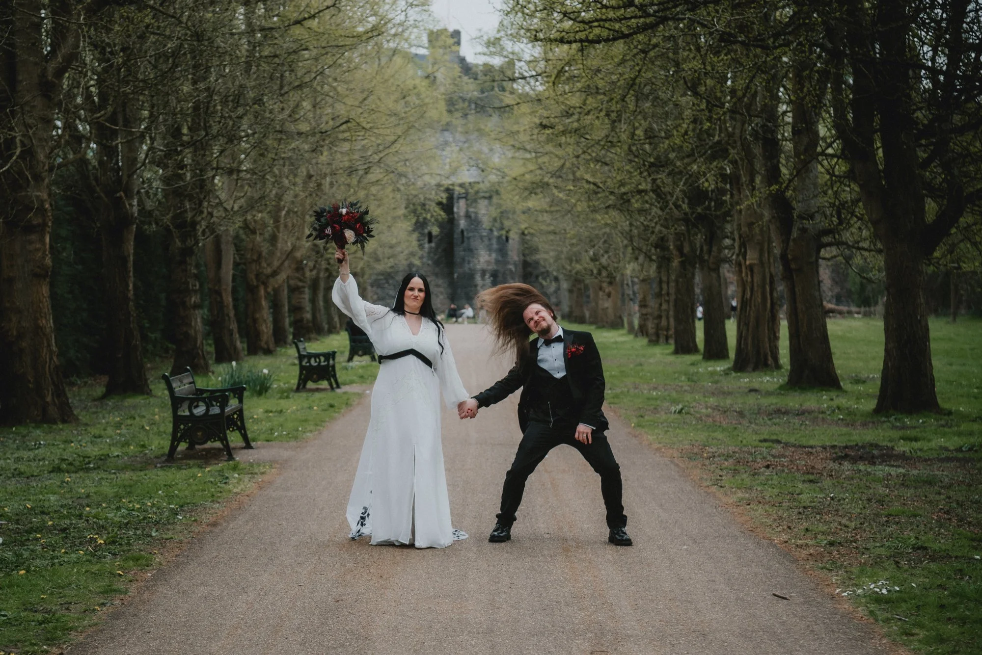 A bride and groom holding hands on a tree-lined path, with the bride holding a bouquet and the groom in a tuxedo, outdoors in a park-like setting.