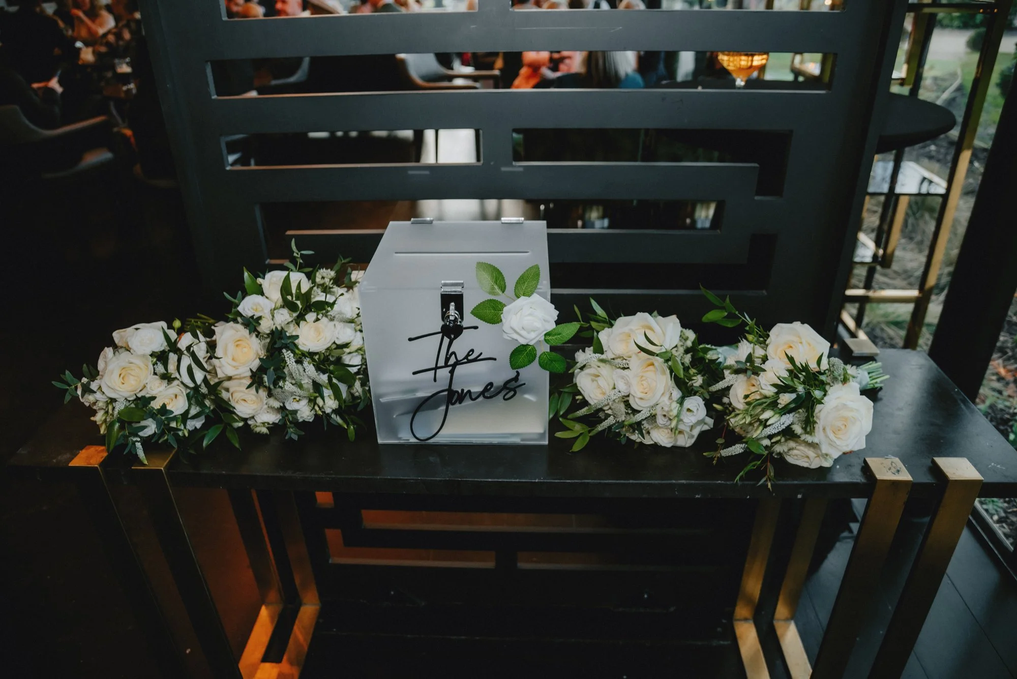 A small table decorated with white roses and greenery, with a white box with black cursive writing that says 'the Jones', surrounded by floral arrangements, in a dimly lit indoor setting with a dark black/gray background.