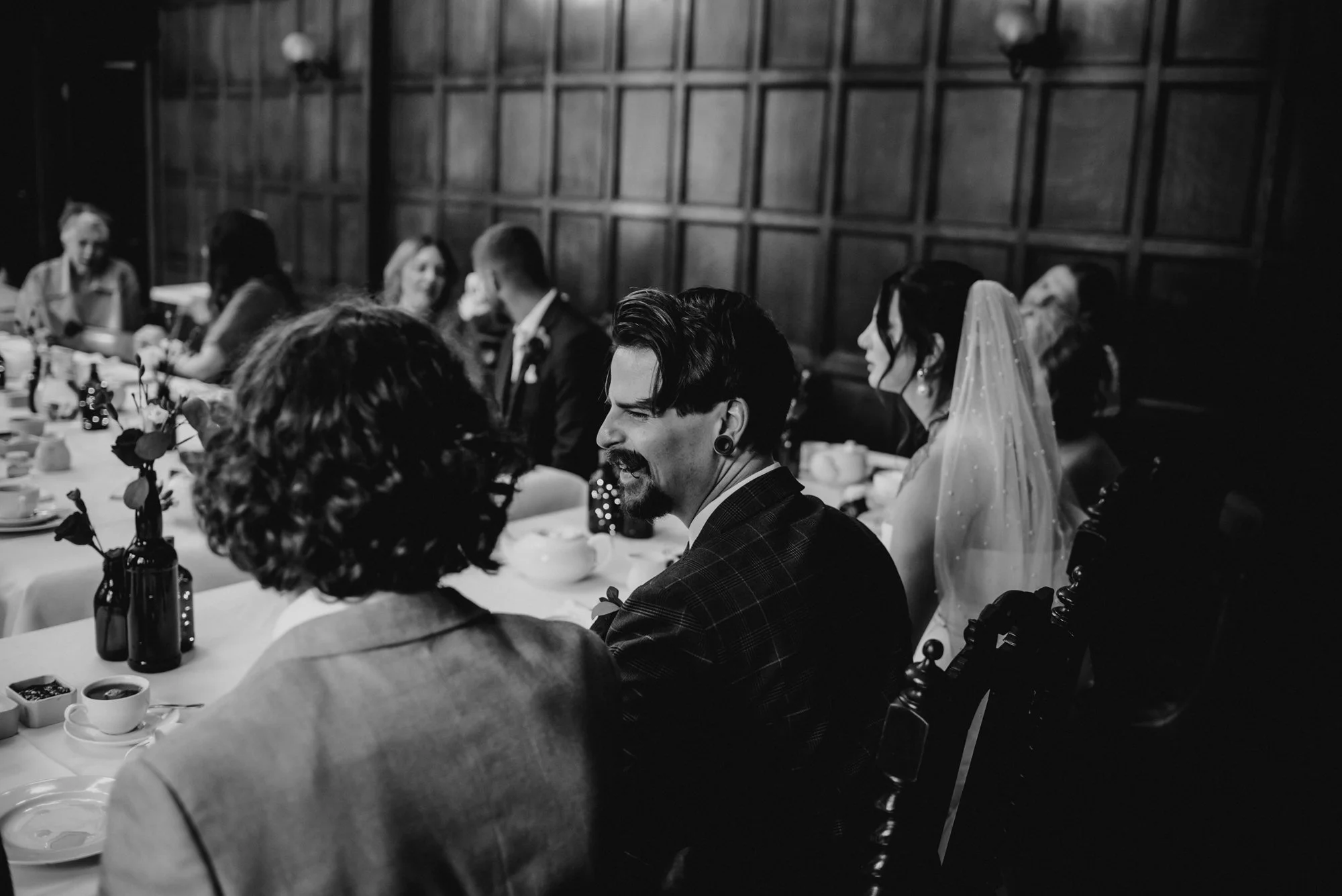 Black and white photo of a wedding reception with guests seated at a long table, including a woman in a veil and a man with a mustache, all seated in a room with dark wooden paneled walls.