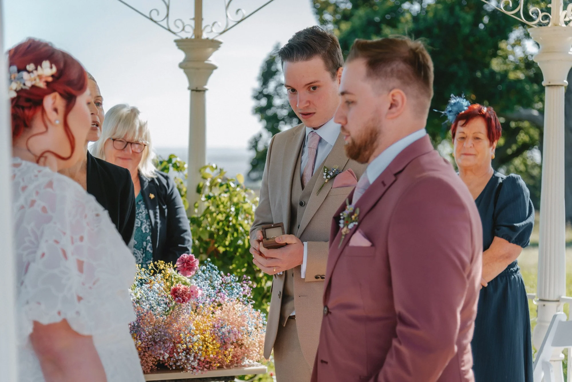 A wedding ceremony with a bride in a white lace dress and red hair, standing in front of two grooms in suits, at an outdoor event under a gazebo with columns, greenery, and a large bouquet of colorful flowers.