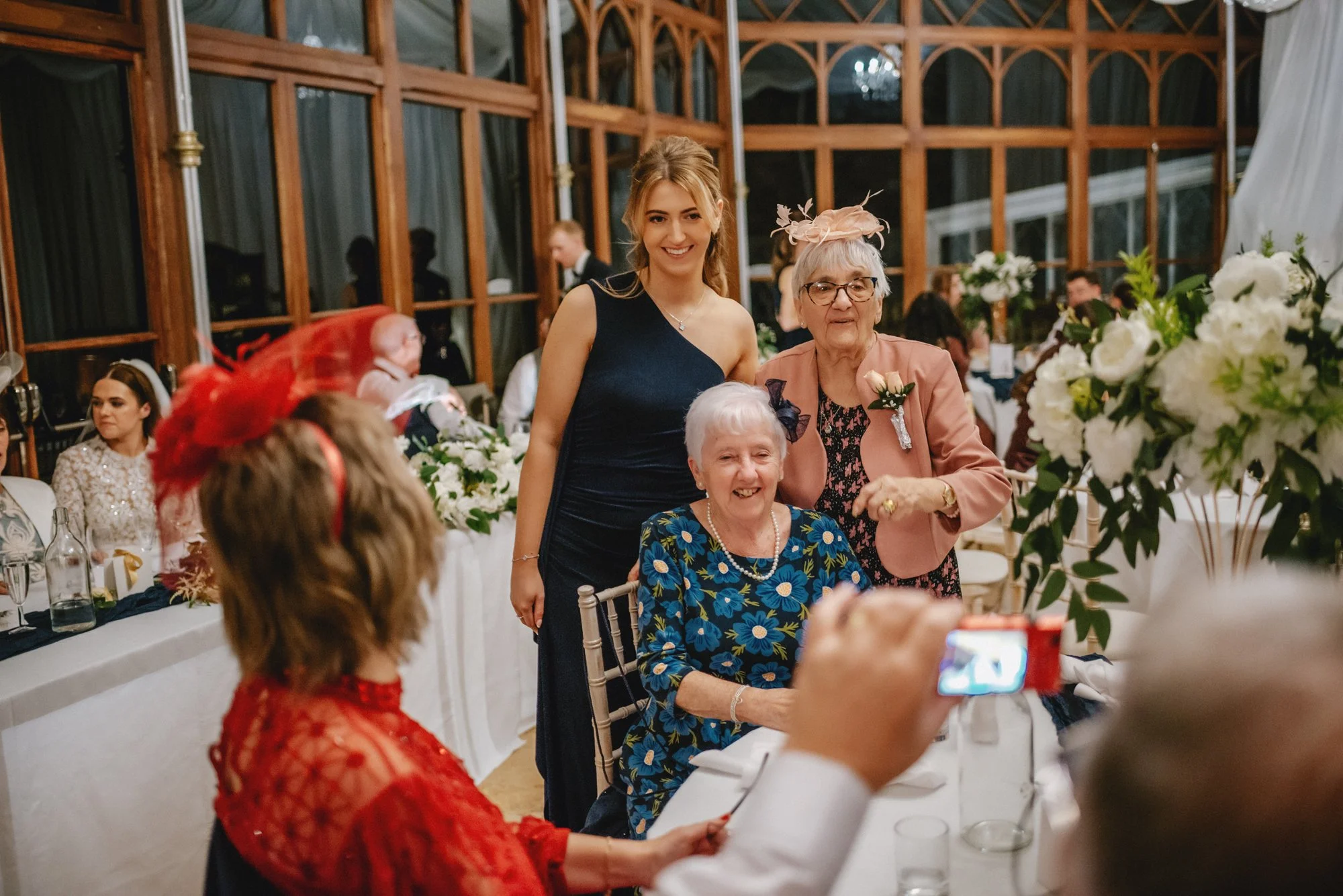 Group of women at a formal celebration, with one elderly woman sitting at a table, two women standing behind her, and others in the background, all dressed in elegant attire, with floral decorations and large windows in the venue.