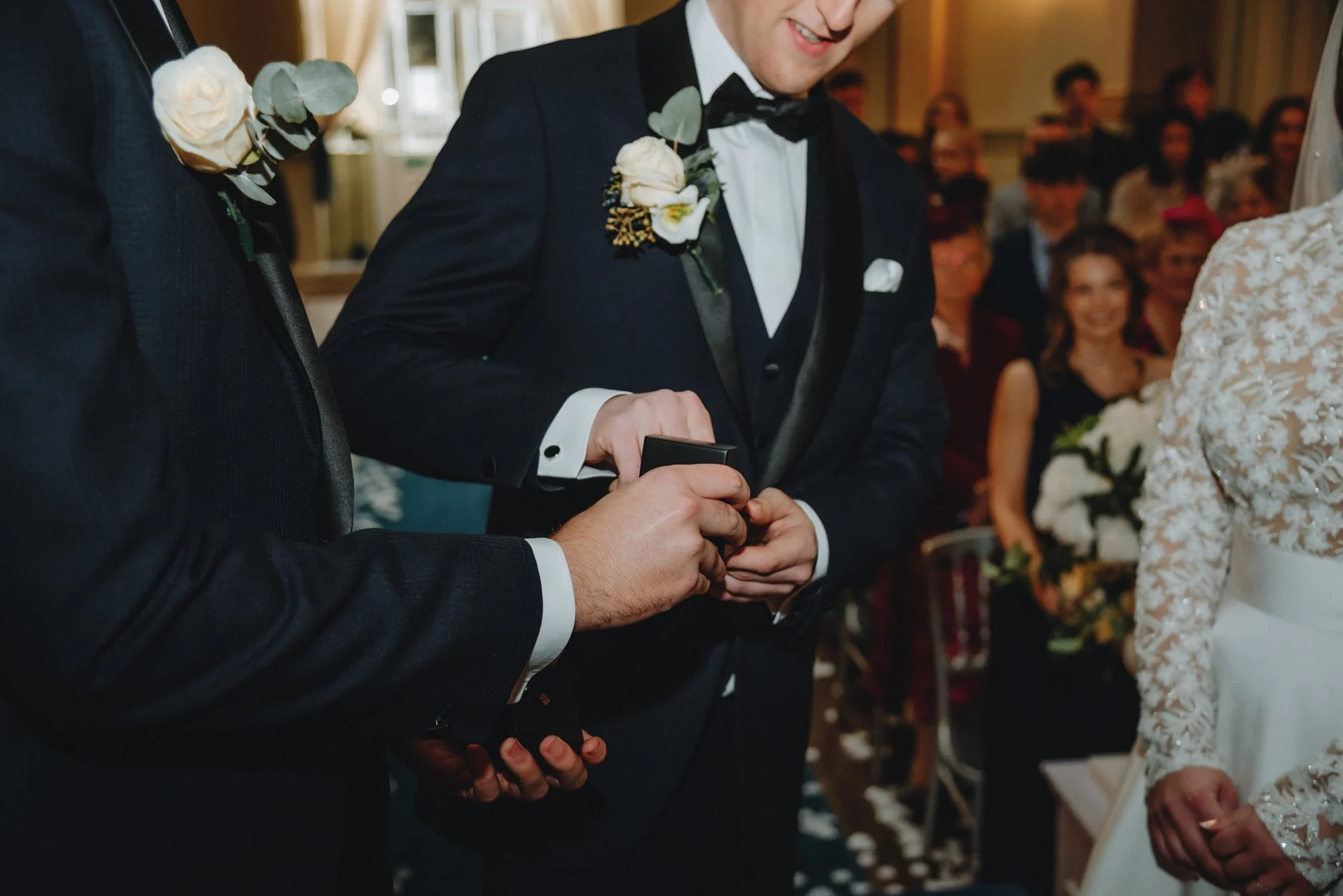 Groom in black tuxedo with boutonniere, holding a ring box during a wedding ceremony, with guests seated in background.
