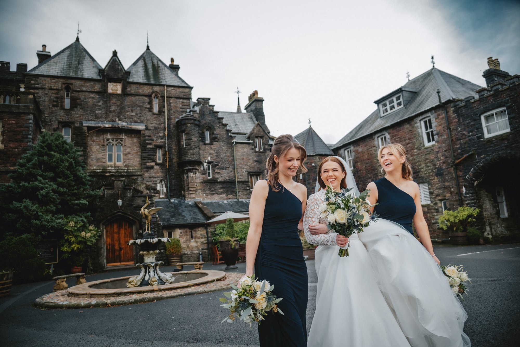 Three women, one in a wedding dress holding a bouquet, and two in navy blue dresses holding bouquets, walking outdoors in front of a historic stone building with a fountain.