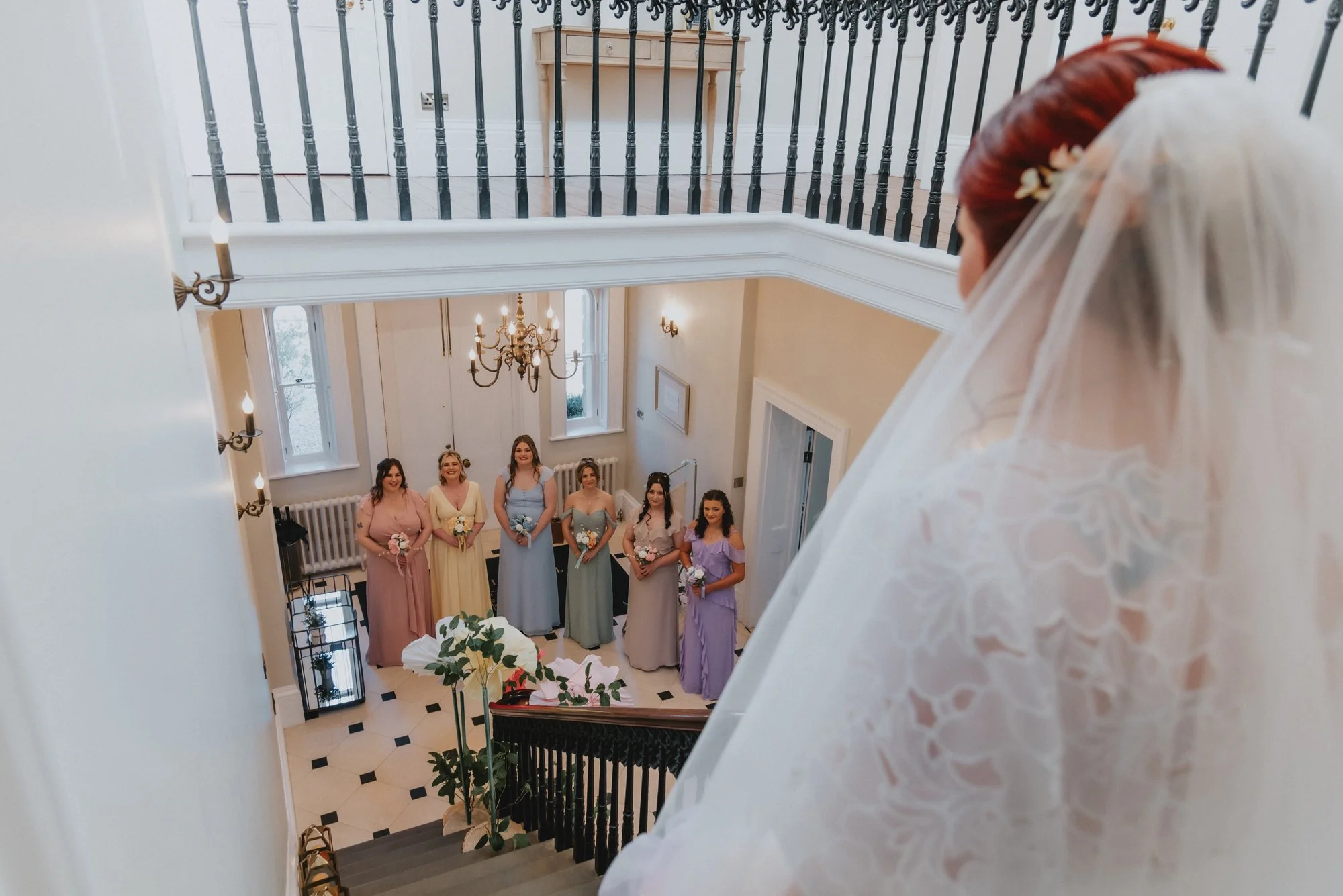 A bride with a veil looks down from a staircase at six bridesmaids standing in a line in a hallway, holding bouquets, wearing pastel colored dresses.