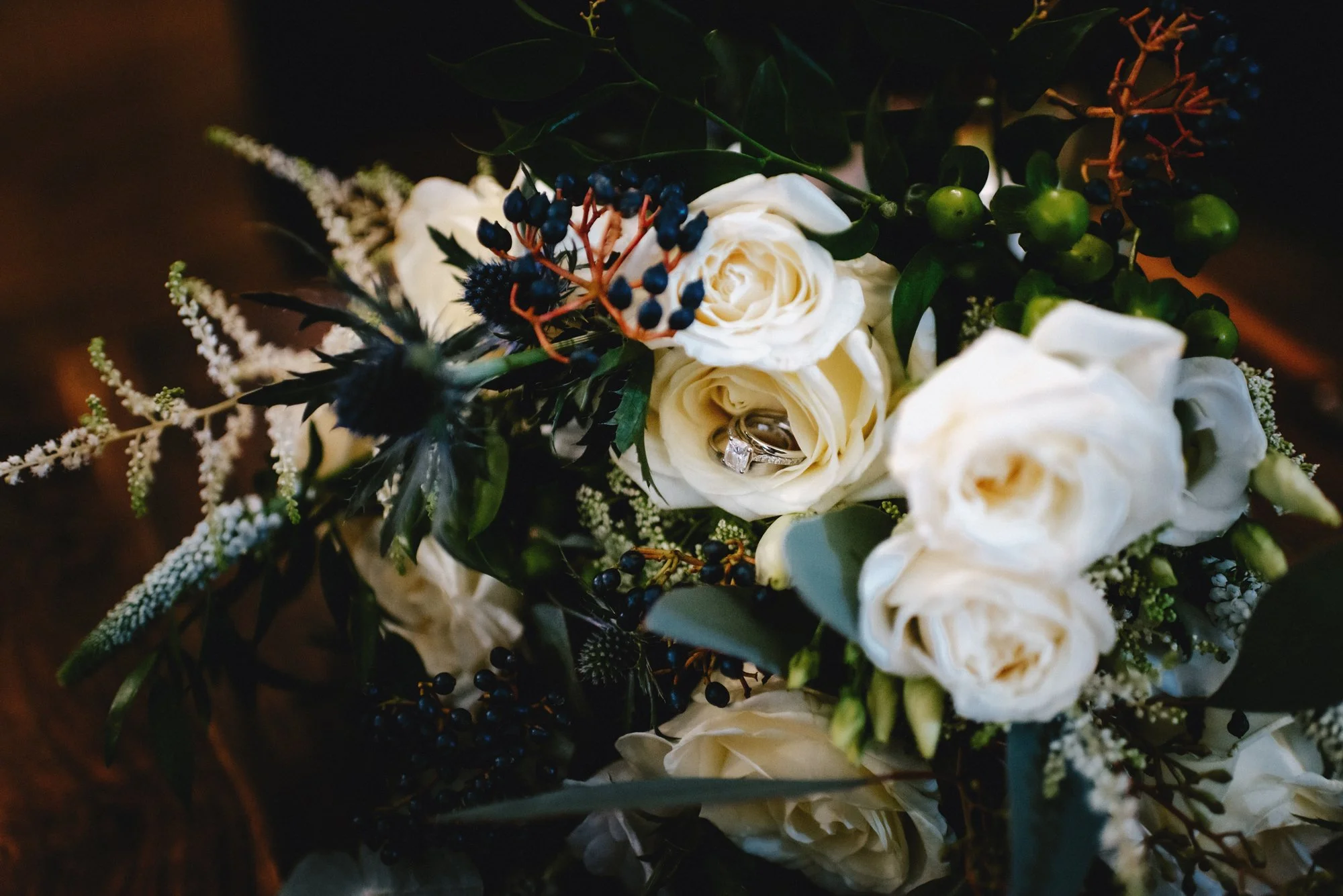 Close-up of a wedding bouquet with white roses, dark blue berries, and greenery, with a diamond engagement ring resting inside one of the roses.