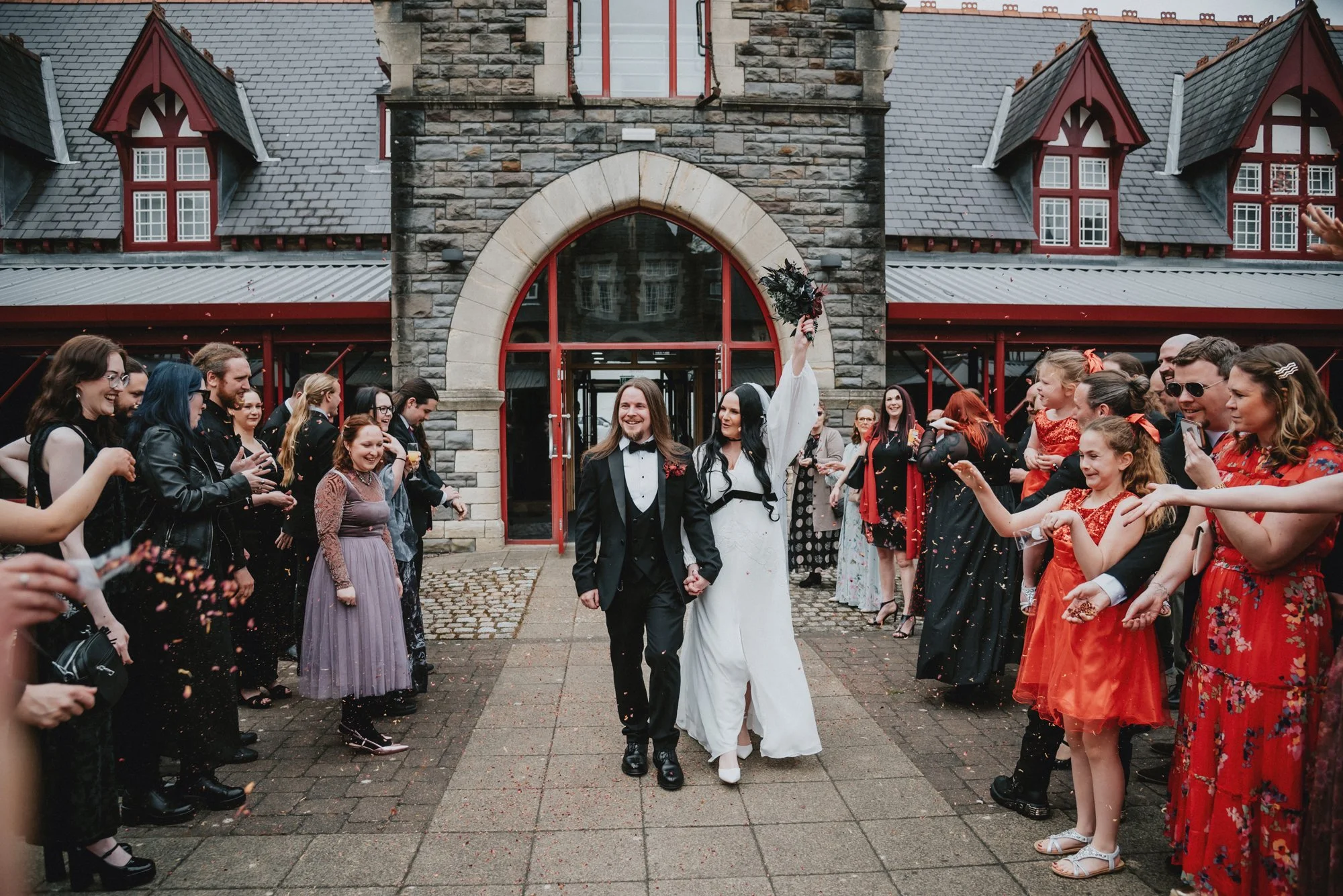 A couple dressed in wedding attire is walking hand in hand through a crowd of celebrating guests outside a stone church with red window frames. The bride, in a white dress, is holding a bouquet high in the air, smiling, while the groom, in a black tu