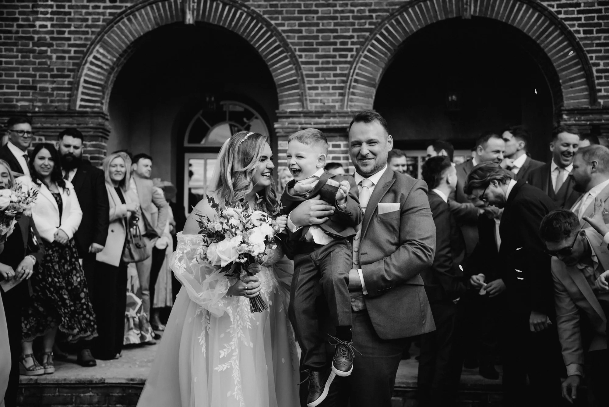 A smiling bride holding a bouquet, a groom carrying a young boy, and a group of wedding guests gathered outside a brick building with arched windows and doors.