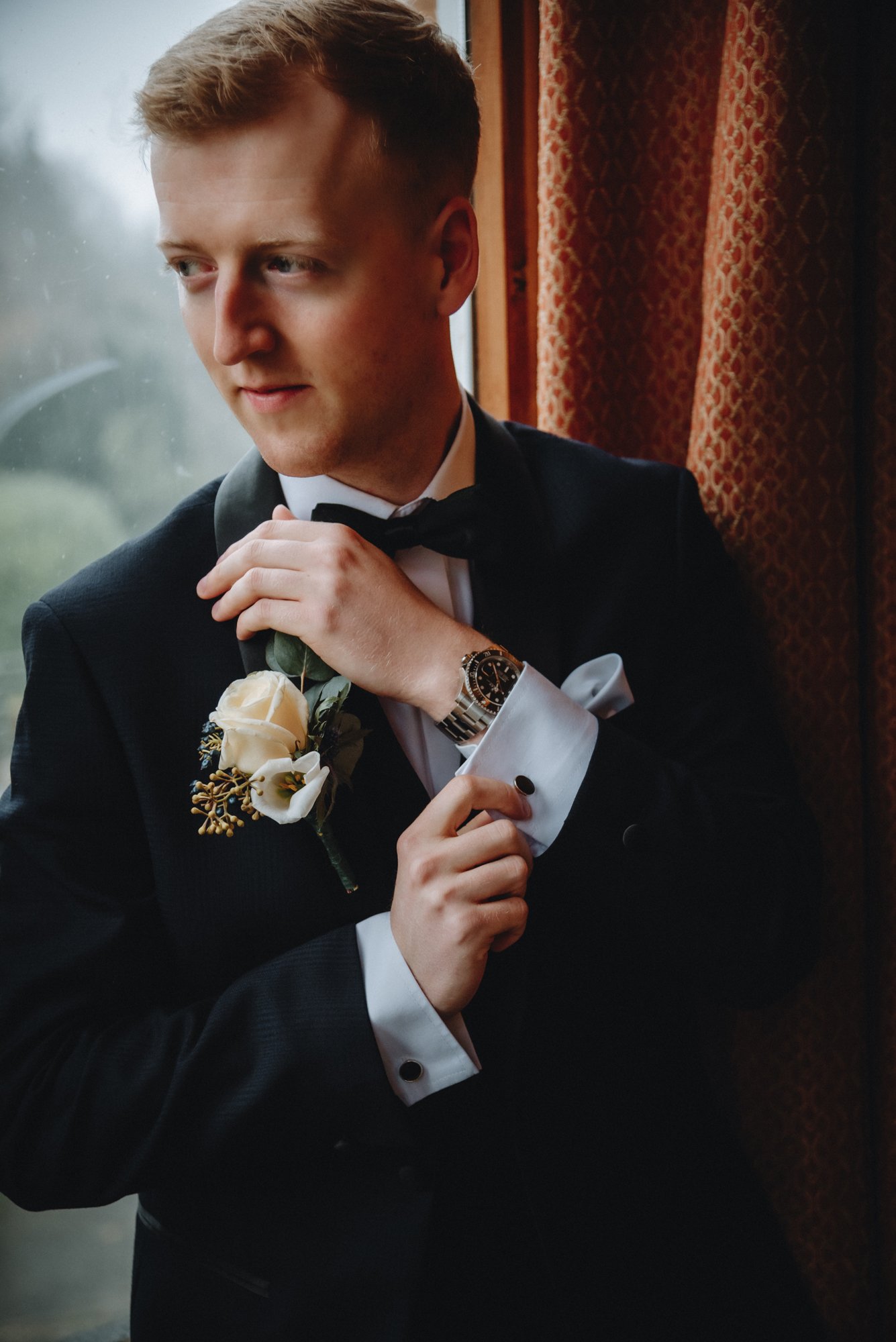 A young man dressed in a tuxedo adjusting his bow tie, standing near a window with patterned curtains, wearing a wristwatch, with a boutonniere on his lapel.