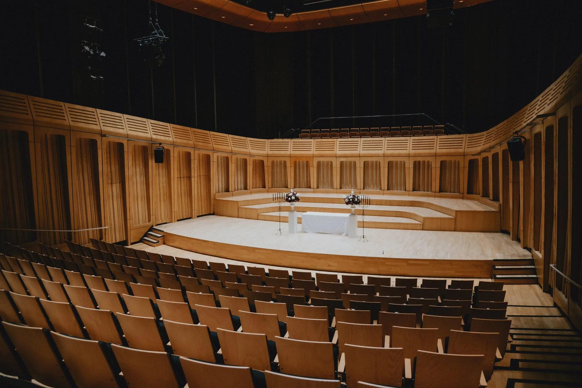 An empty theater stage with wooden paneling, decorated with flowers and candelabras, set up for a wedding or event, with rows of chairs facing the stage.