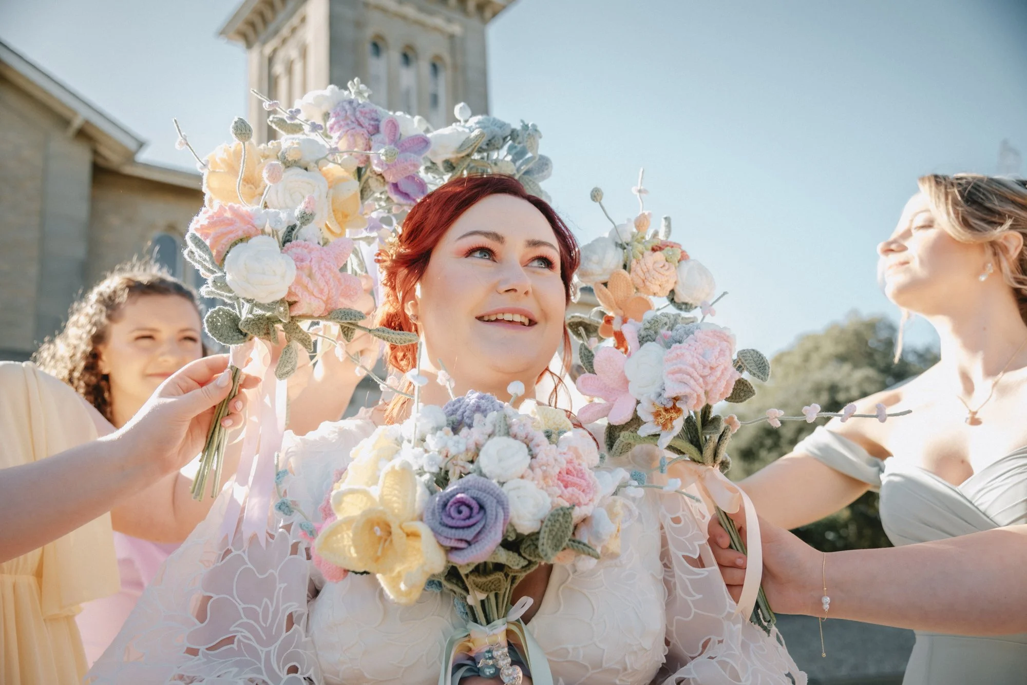 A bride with red hair surrounded by women holding floral bouquets during an outdoor wedding ceremony