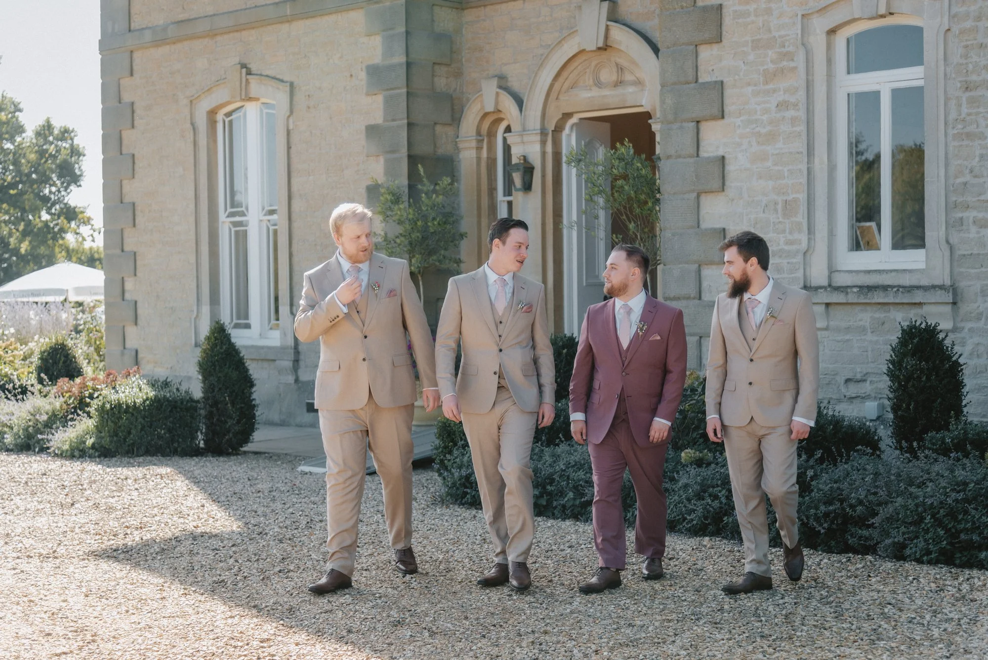 Four men in suits walking outdoors near a stone building, engaged in conversation.