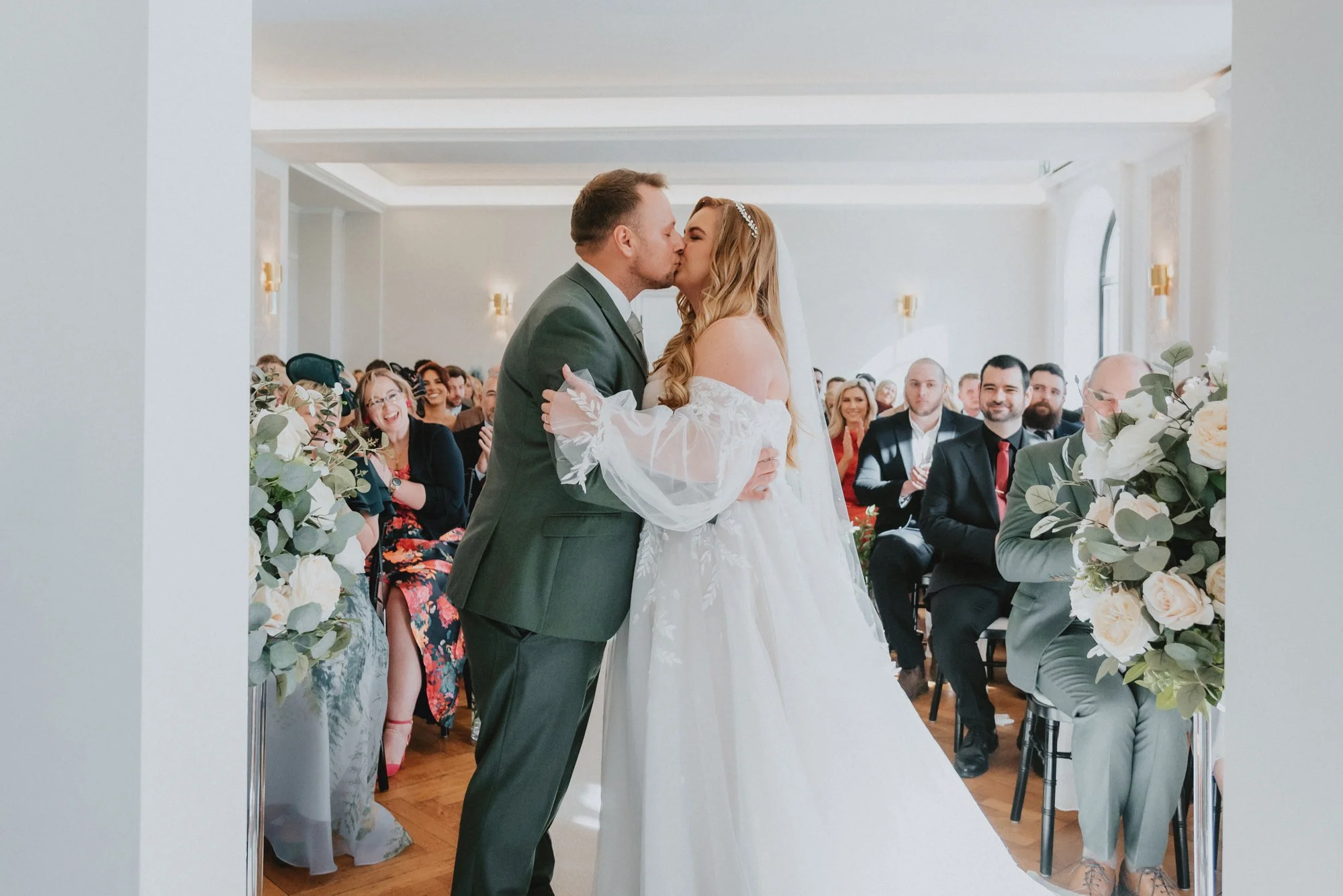 A bride and groom share a kiss at their wedding ceremony, surrounded by seated guests in a decorated venue.