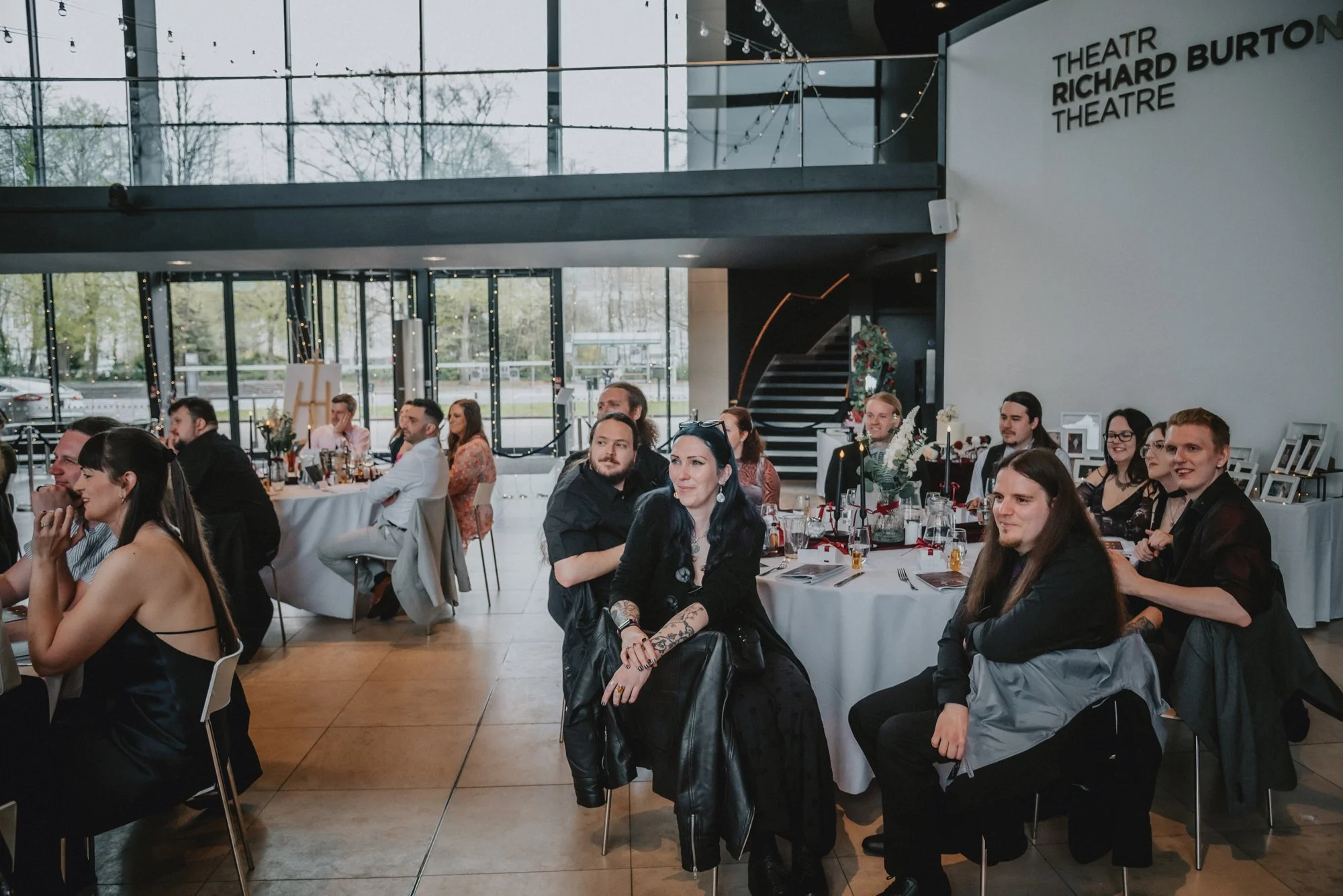 People sitting at decorated tables in a modern event space with large windows, during a gathering or celebration.