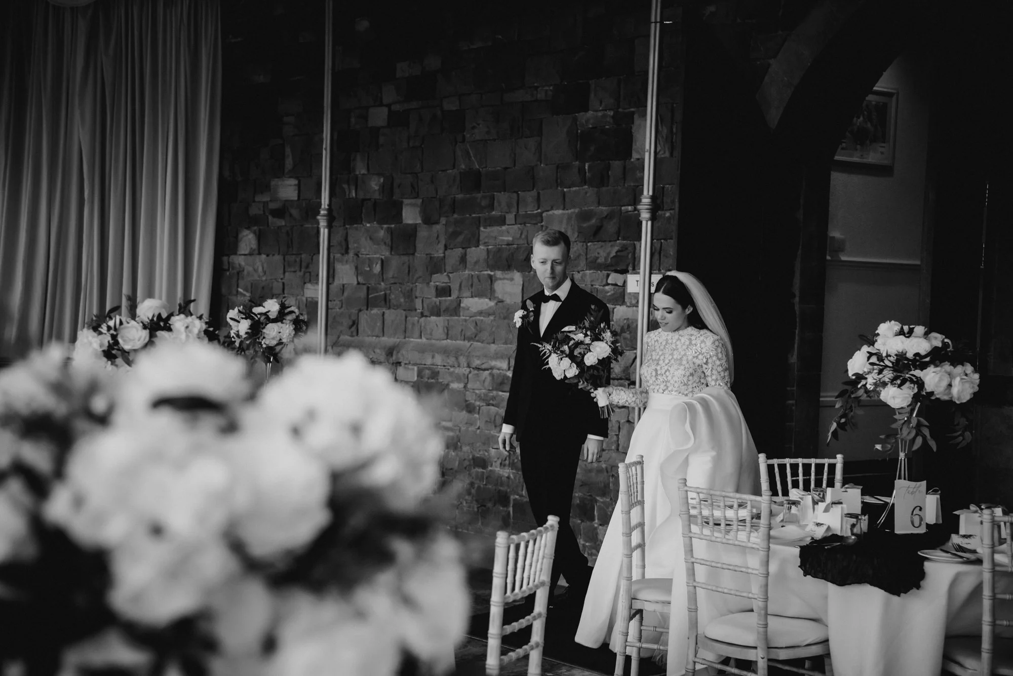 Black and white photo of a bride and groom standing together at a wedding reception. The bride wears a long-sleeved lace wedding gown and veil, holding a bouquet. The groom wears a tuxedo with a bow tie, holding a bouquet. They are in a decorated ven