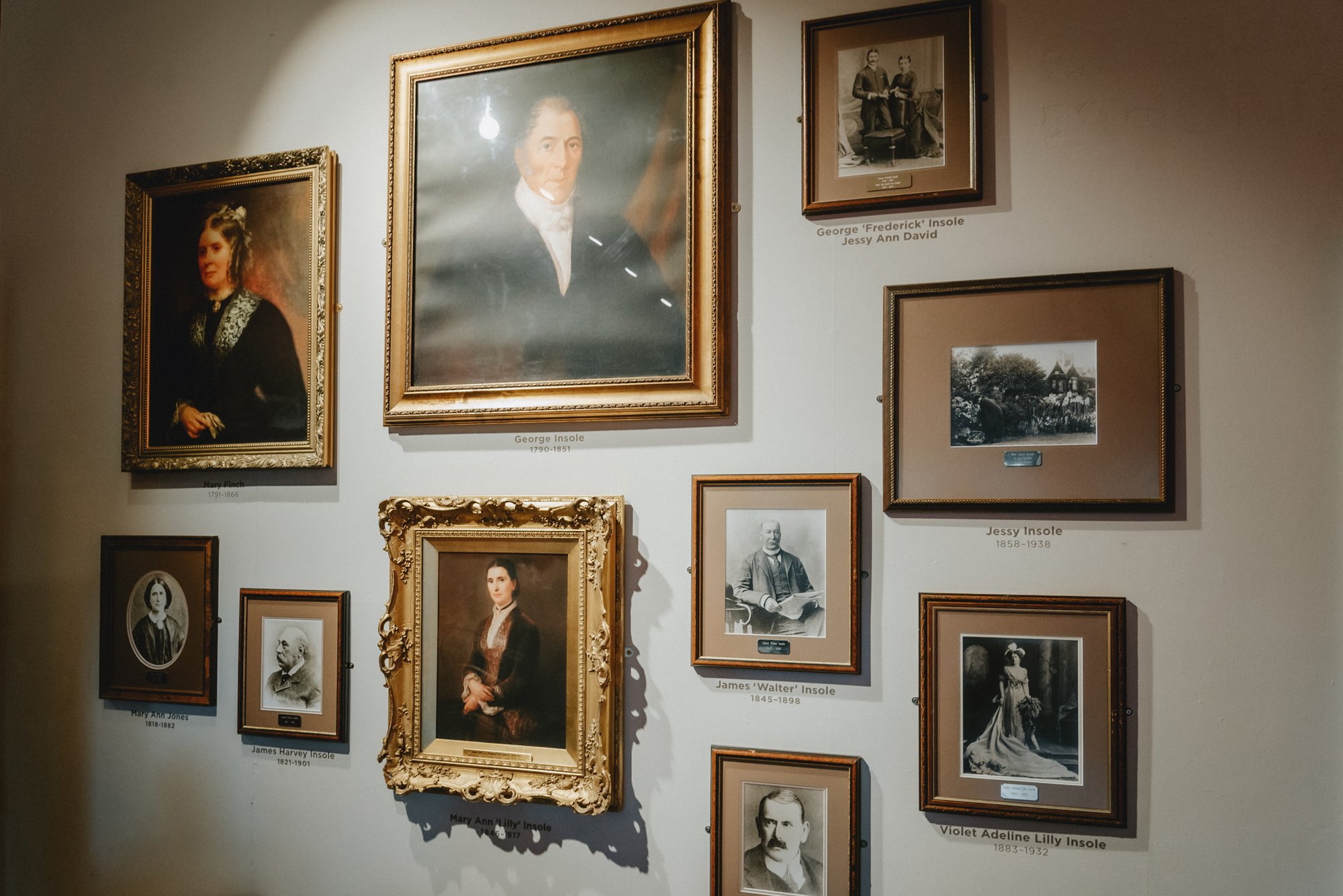 A wall displaying various framed portraits of people, including men and women, some in black-and-white photographs and others in painted portraits, with labeled names and birth-death years.