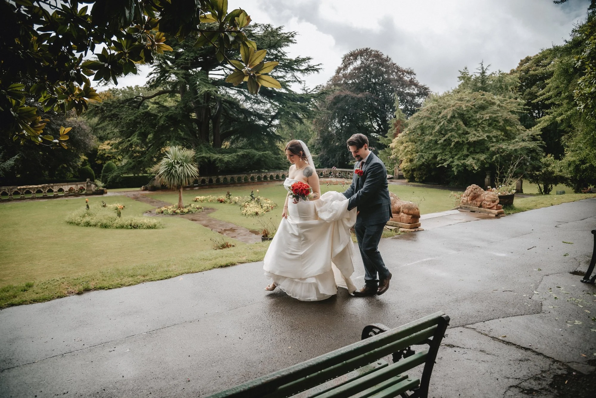 A bride and groom outside on a rainy day, with the groom helping the bride walk. The bride wears a white wedding dress and holds a bouquet of red roses; the groom is in a dark suit with a red boutonniere. They are on a paved pathway surrounded by a l