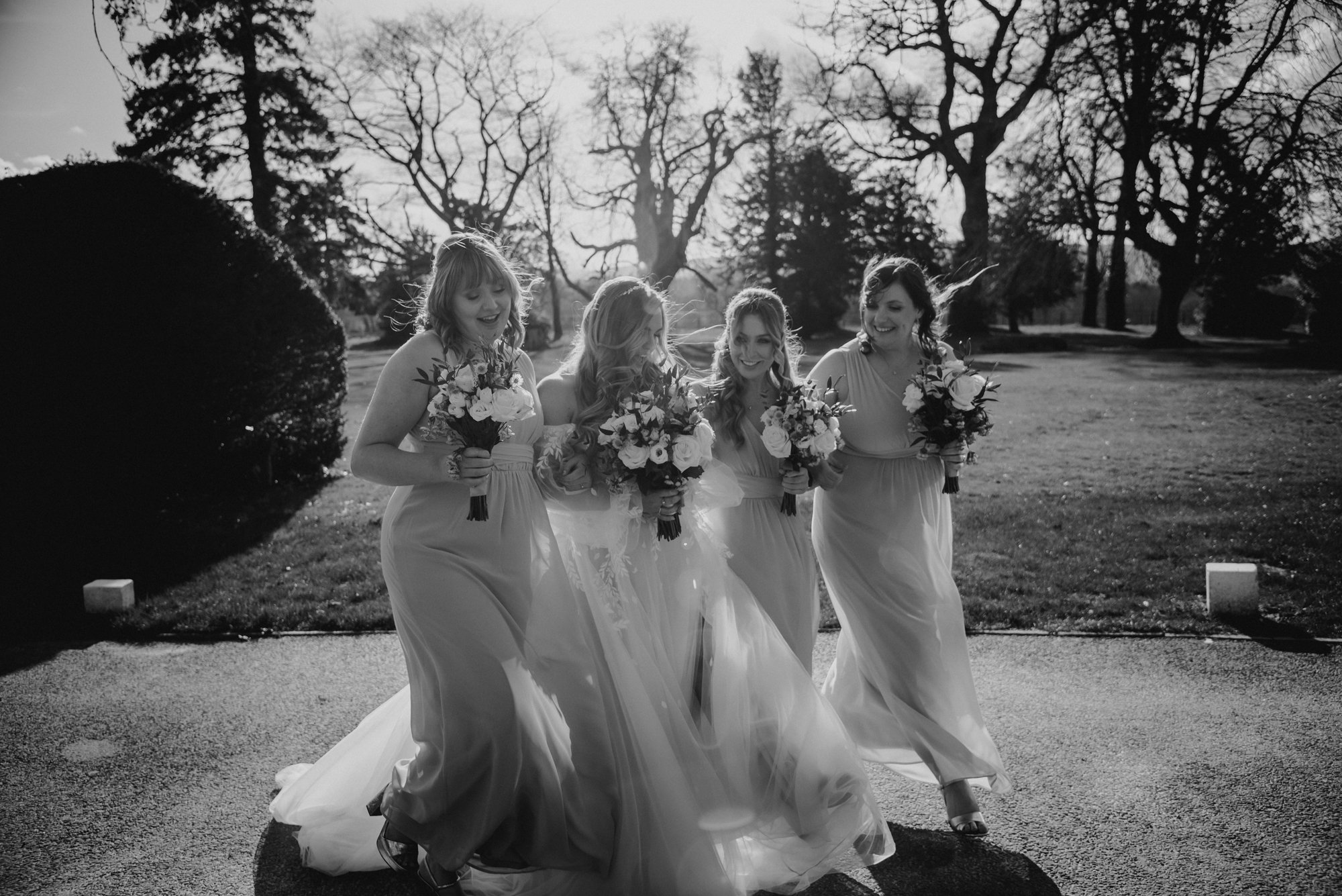 Four women in wedding dresses walking outdoors, holding bouquets of flowers, smiling and enjoying themselves with leafless trees and a grassy area in the background.