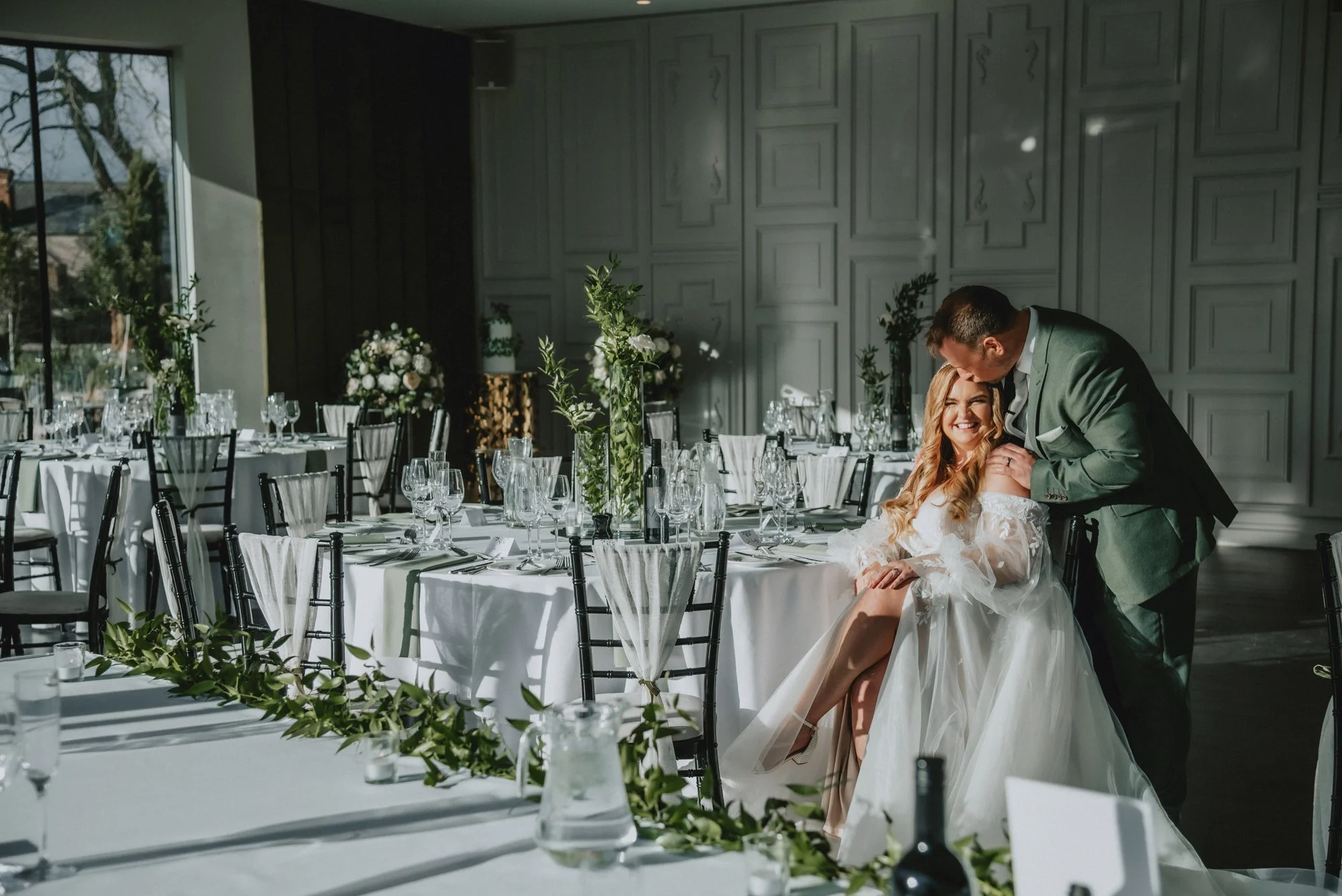 A bride and groom sharing a loving moment at their wedding reception in a decorated banquet hall with sunlight streaming through large windows.
