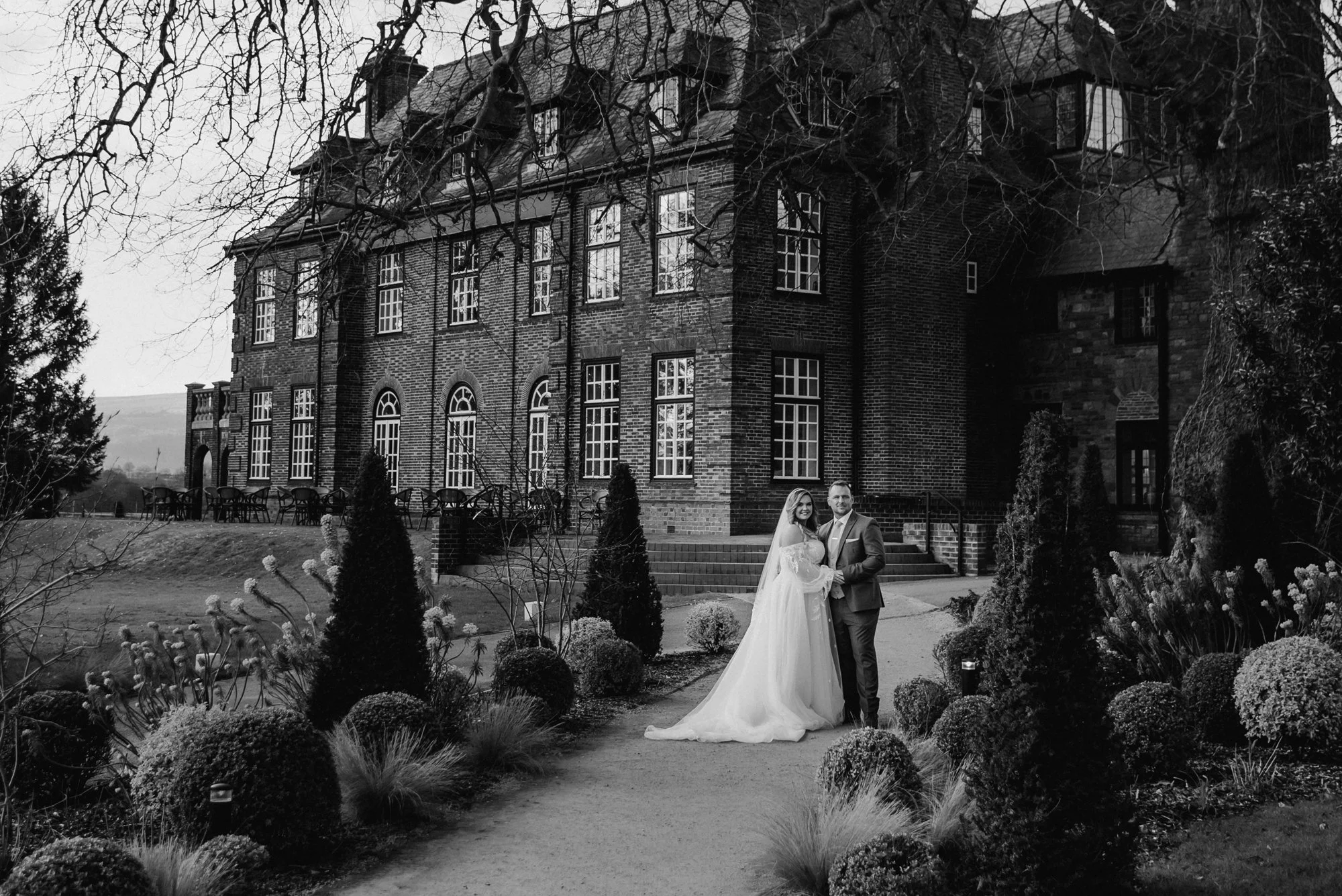 A bride and groom standing hand in hand in front of a large brick mansion surrounded by well-manicured bushes and trees, in a wedding photo setting.