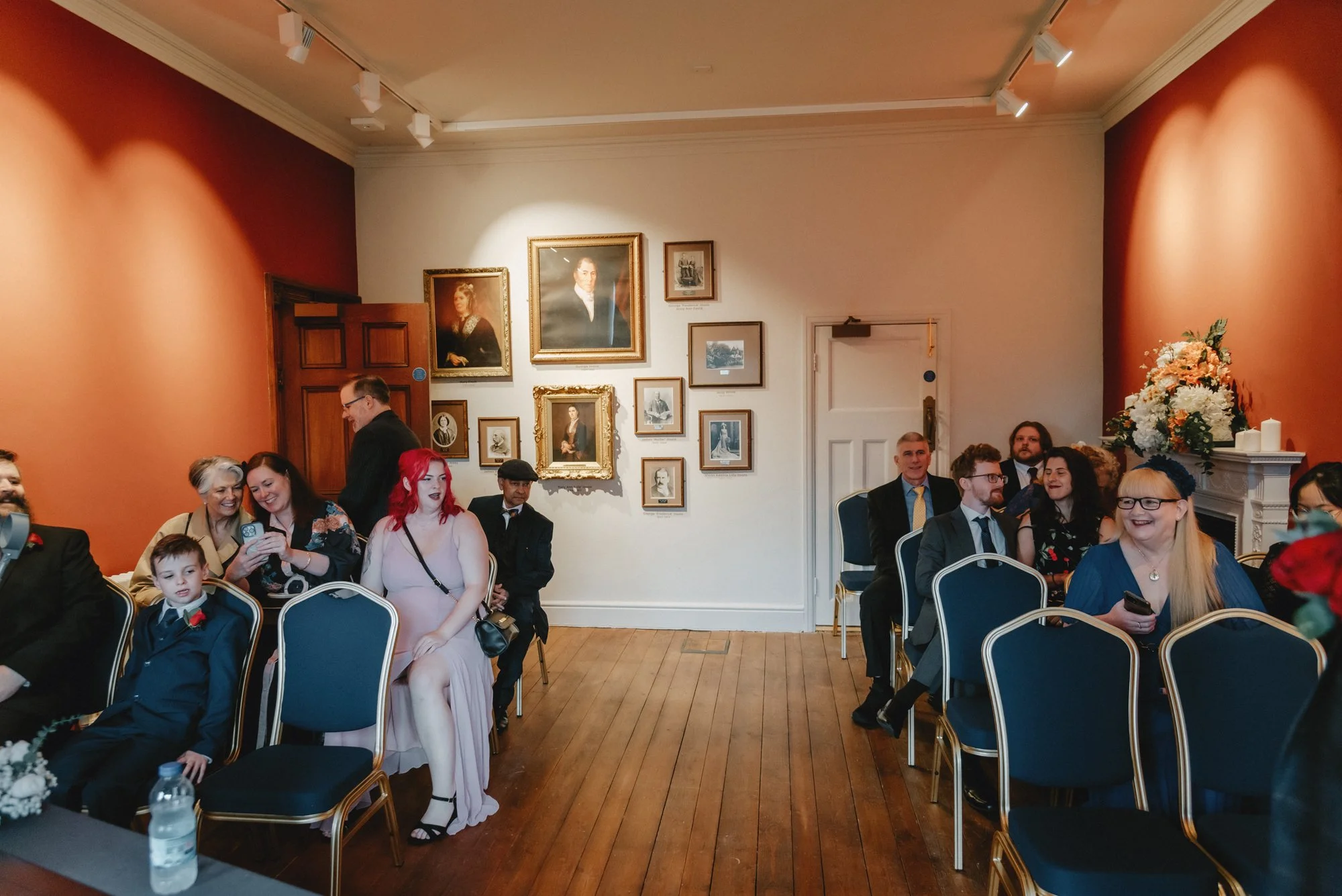 People seated in an elegantly decorated room with framed portraits on the wall, attending an event or gathering.
