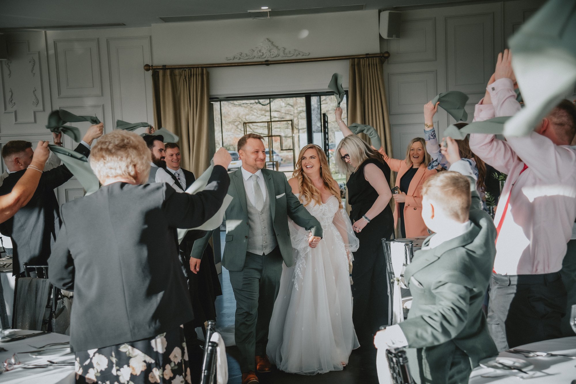 A bride and groom walking hand-in-hand into a celebration with friends and family, some holding napkins or cloths, in a well-lit decorated venue.