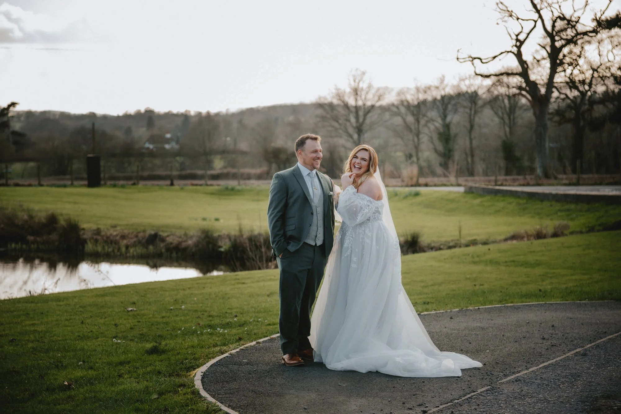 A bride and groom standing outdoors on a circular paved area, smiling and laughing, with a pond and trees in the background during late afternoon or early evening.