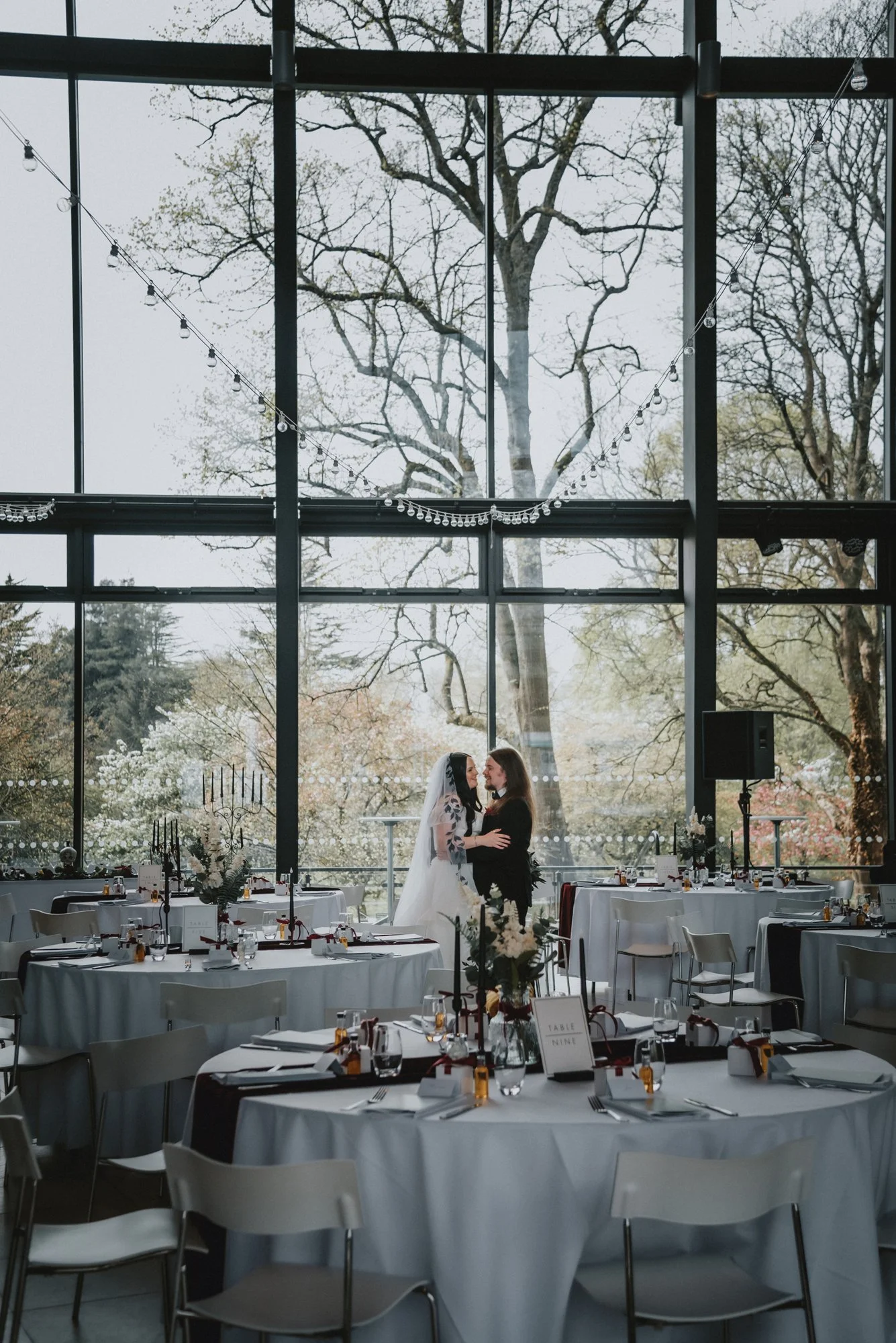 Wedding reception with two women, one in a white dress and veil, and the other in a black dress, standing close together inside a glass-walled venue, with trees visible outside, decorated tables, and string lights.
