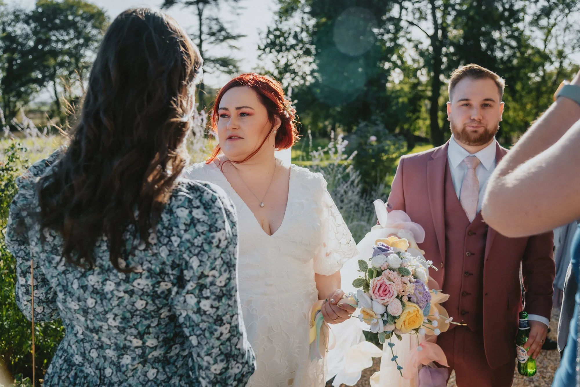 A wedding scene outdoors with women and a man, one woman holding a bouquet of flowers, dressed in formal attire, with trees and sunlight in the background.