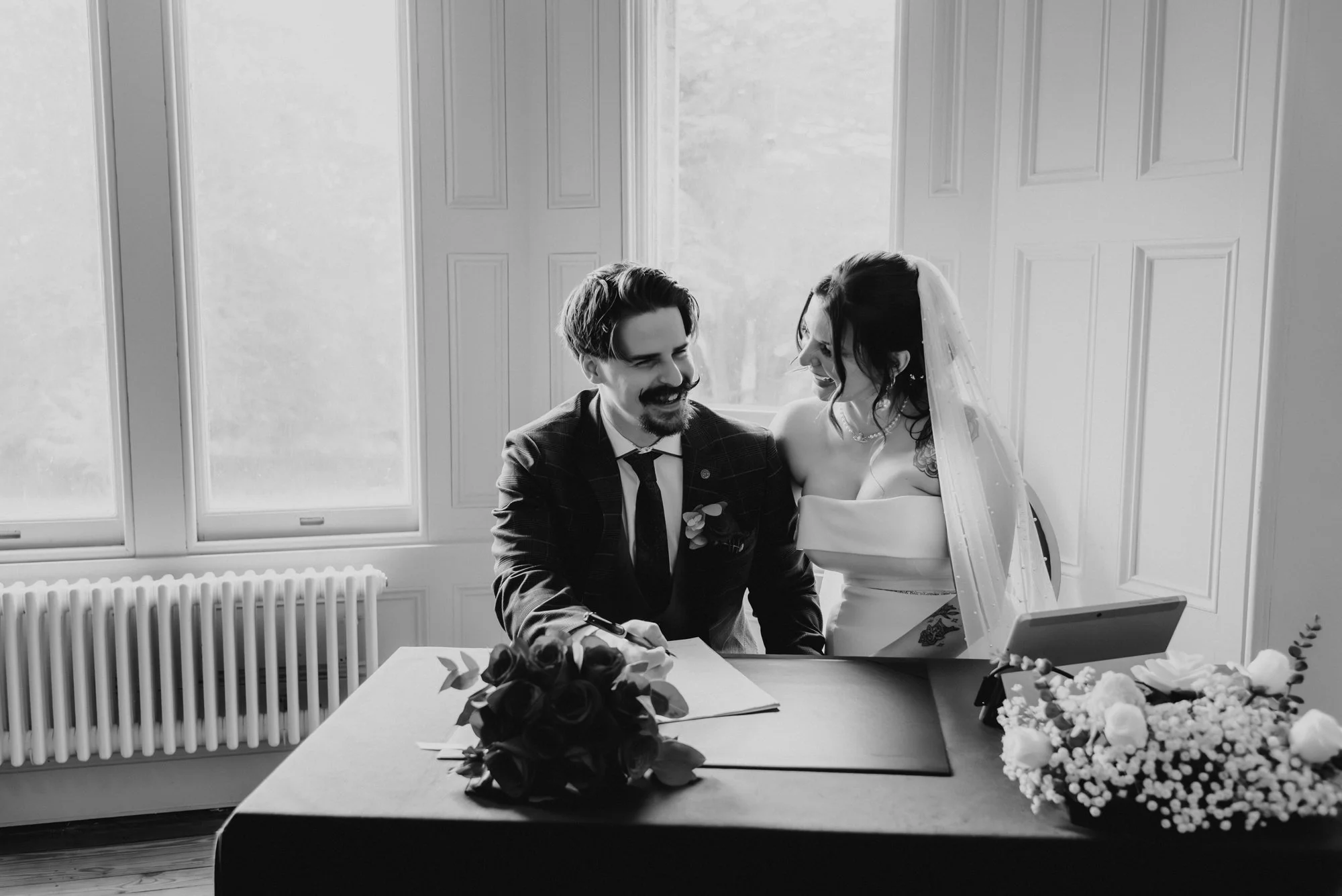 A black and white photo of a bride and groom sitting at a table, smiling and looking at each other. The bride is wearing a wedding dress and a veil, with tattoos visible on her shoulder. The groom is dressed in a suit, signing a book with a bouquet o