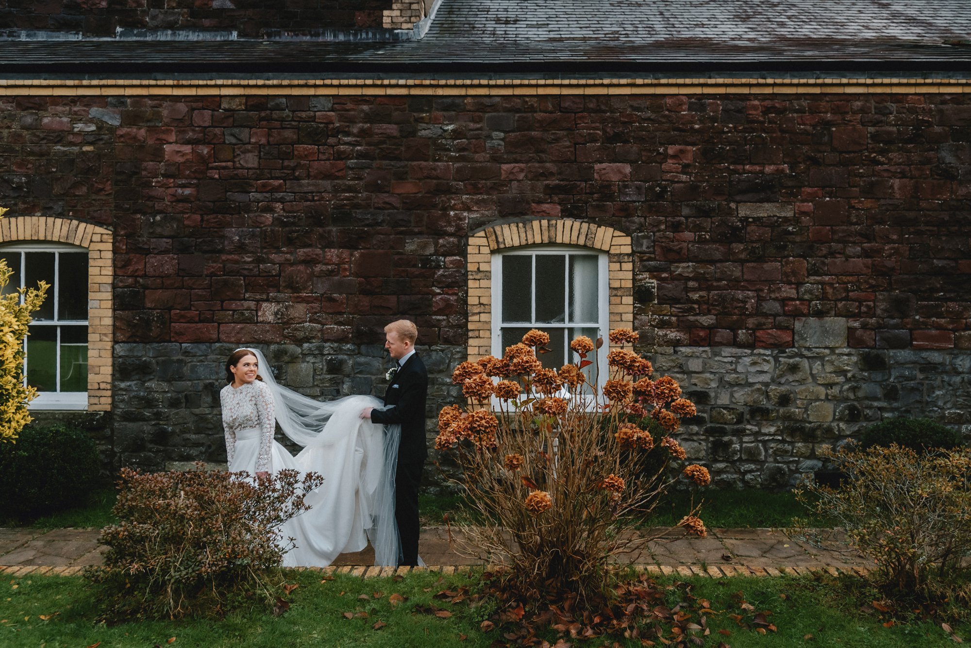 A bride and groom on their wedding day outdoors, with the bride smiling at the groom, who is holding her white wedding dress and veil, in front of a brick and stone building with two windows, surrounded by autumnal bushes and grass.