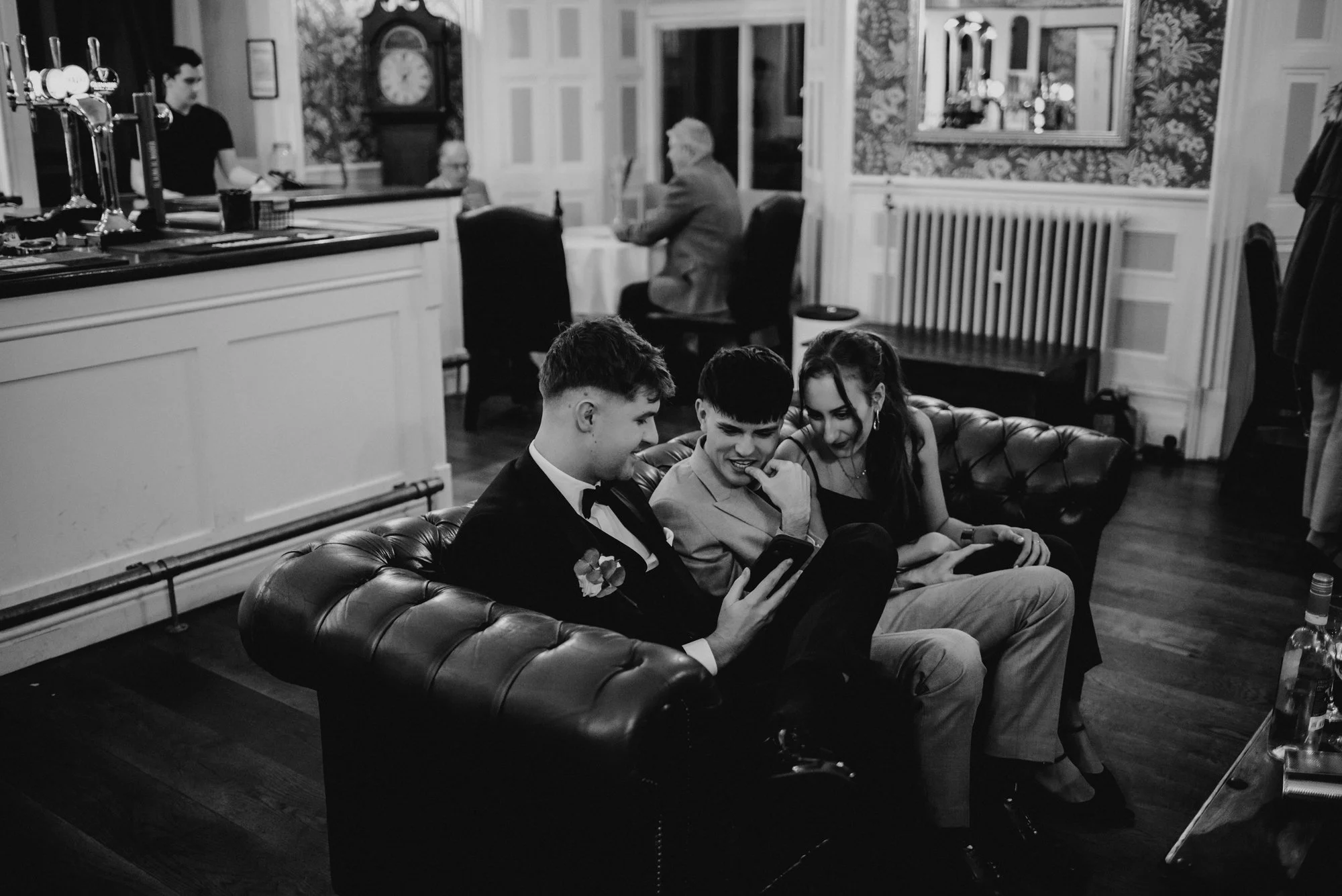 Three young adults sitting on a leather couch, looking at a phone together, in a restaurant or bar setting, in black and white.