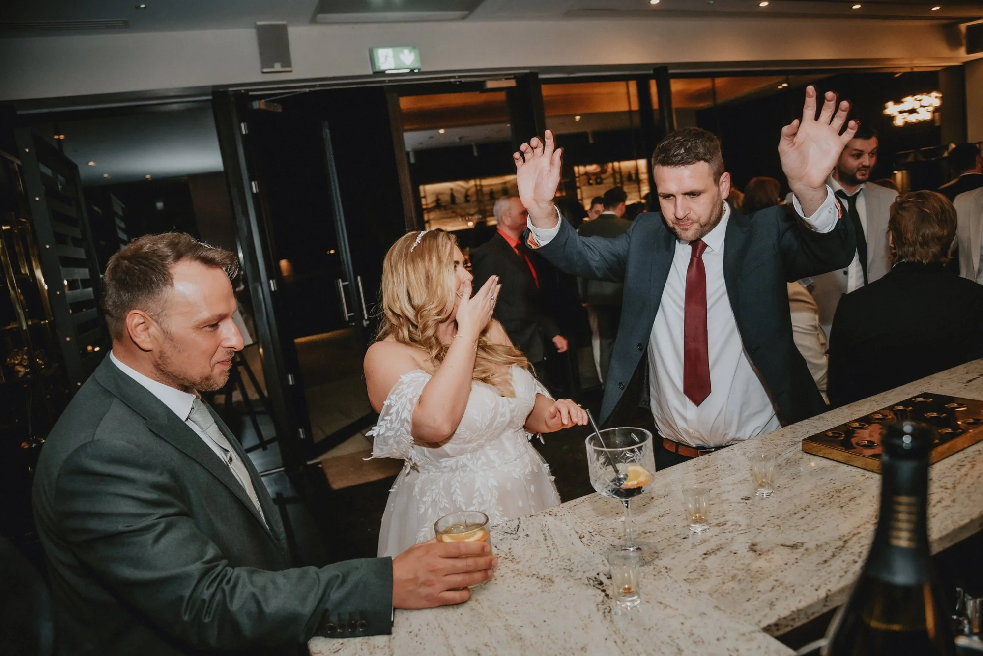 A man in a suit and tie dancing with a raised arm at a party, with a woman in a white dress covering her mouth with her hand and holding a drink, and another man in a suit sitting at a bar counter holding a drink, in an indoor event space with severa