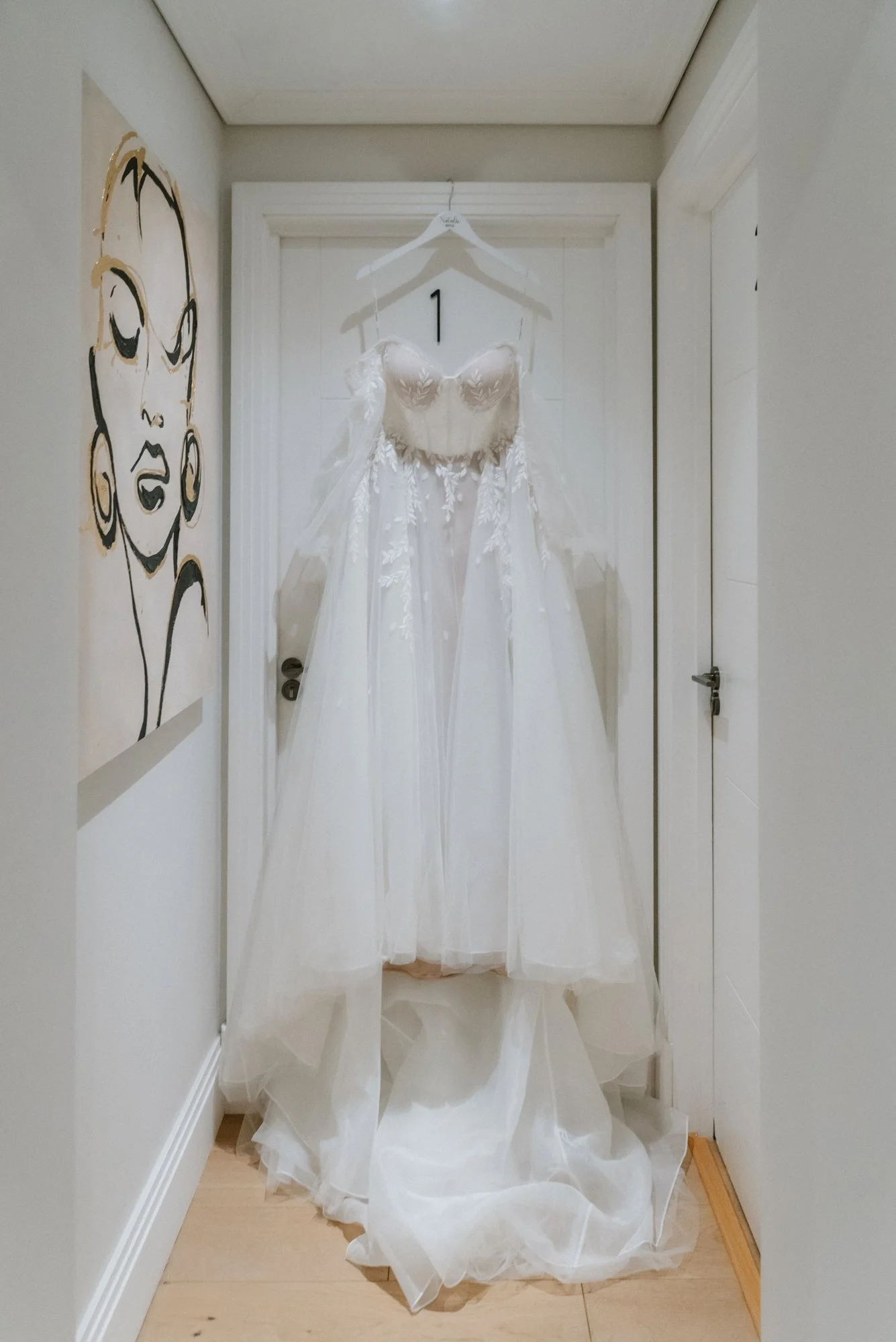 A wedding dress hanging on a hanger on a door with the number 1, in a small hallway with artwork on the wall.