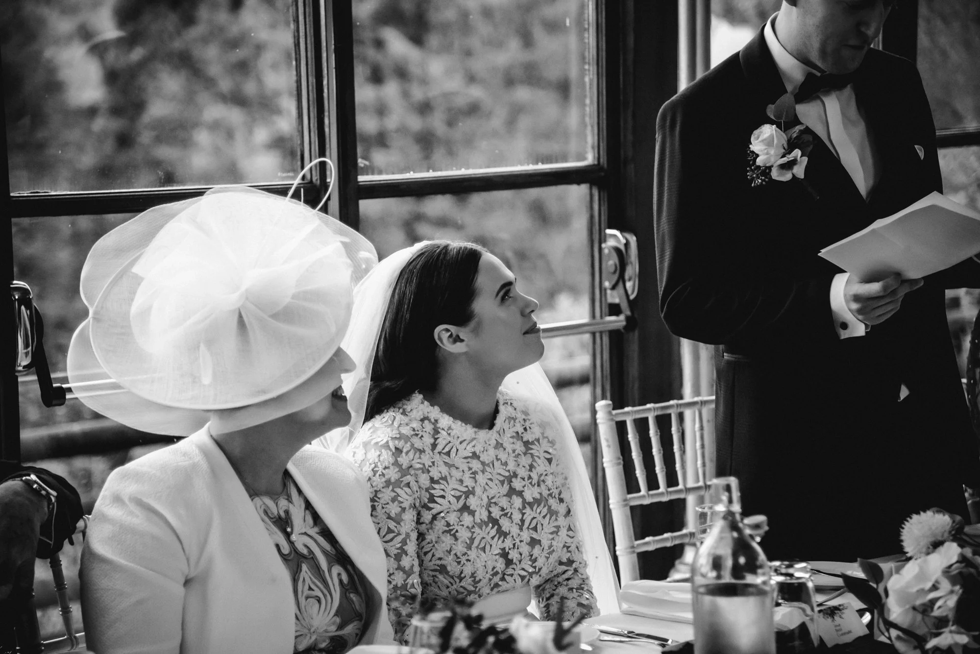 A black-and-white photo of a wedding reception, featuring a woman in a hat and a woman in a bridal gown sitting at a table, while a man in a tuxedo reads from a paper.