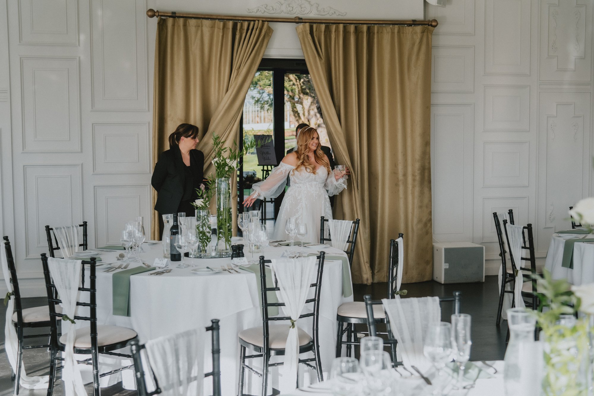 A bride in a white wedding dress standing near the window, holding a glass, with a woman in black standing nearby in a decorated reception room.