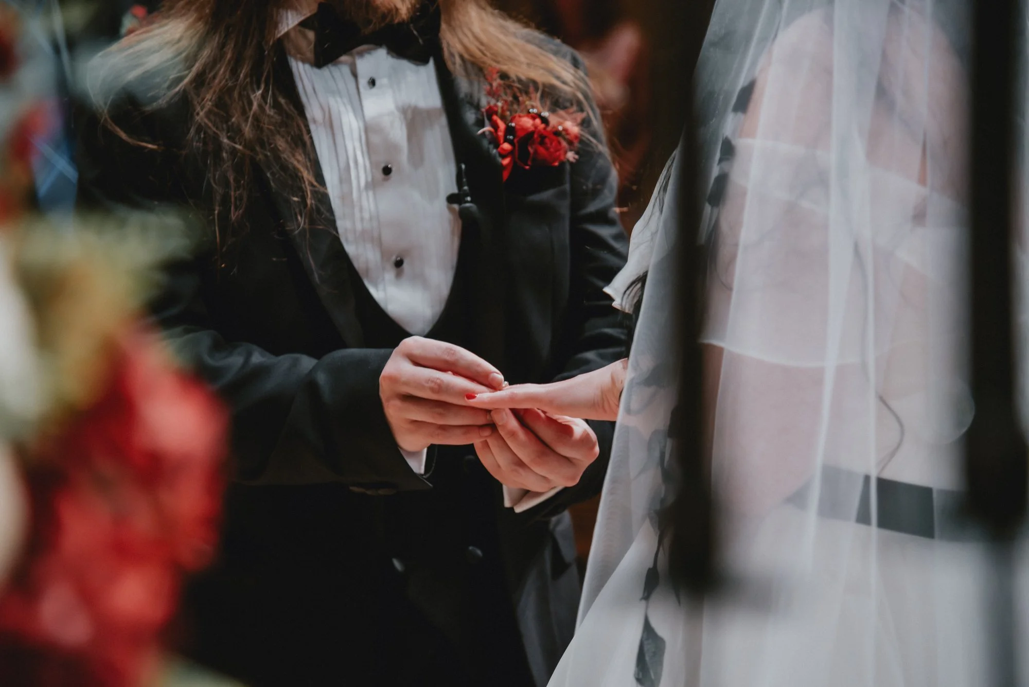 A person in a tuxedo with a red floral boutonniere exchanges wedding rings with another person wearing a wedding dress.