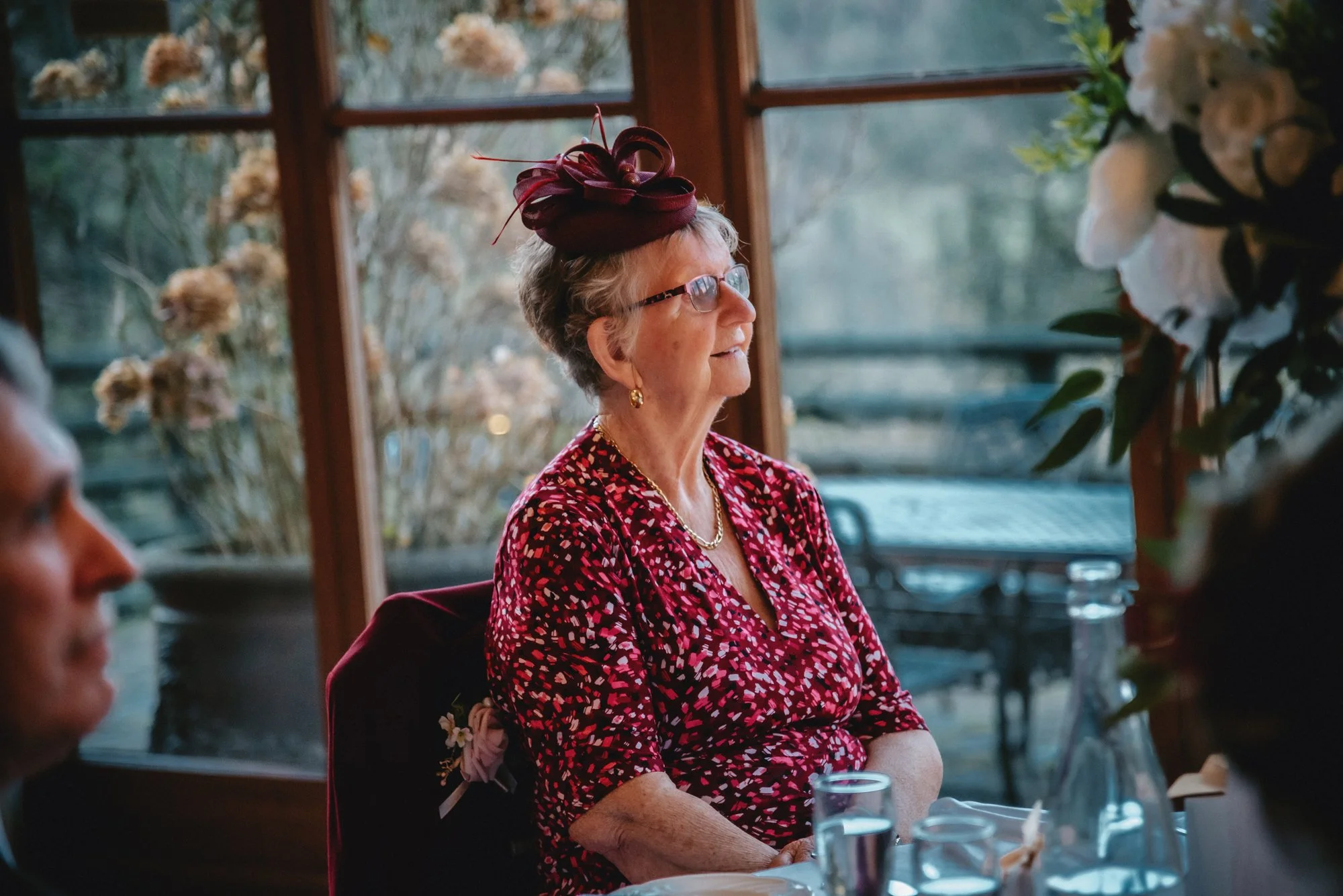 An elderly woman dressed in a red floral dress and a maroon fascinator hat, sitting at a table during a daytime gathering with large windows and floral arrangements in the background.