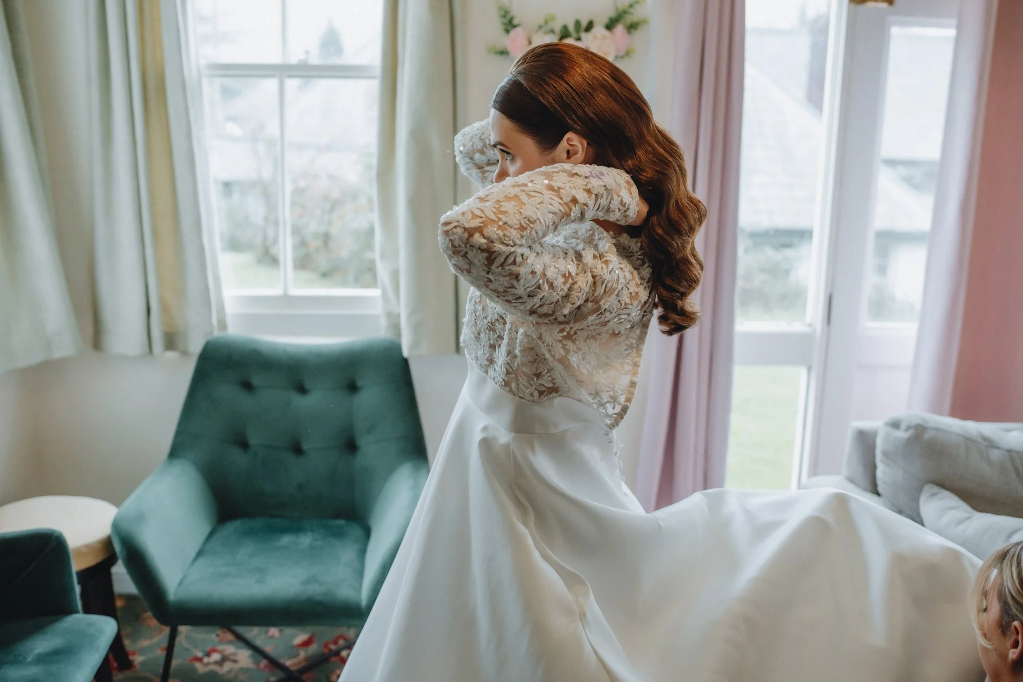 A woman in a wedding dress adjusting her hair in a room with large windows and green chairs.