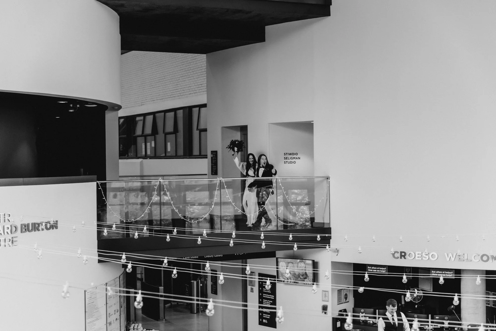 Black and white photo of two women on a balcony in a modern indoor space, one holding a bouquet, with decorative lighting and signage in the background.