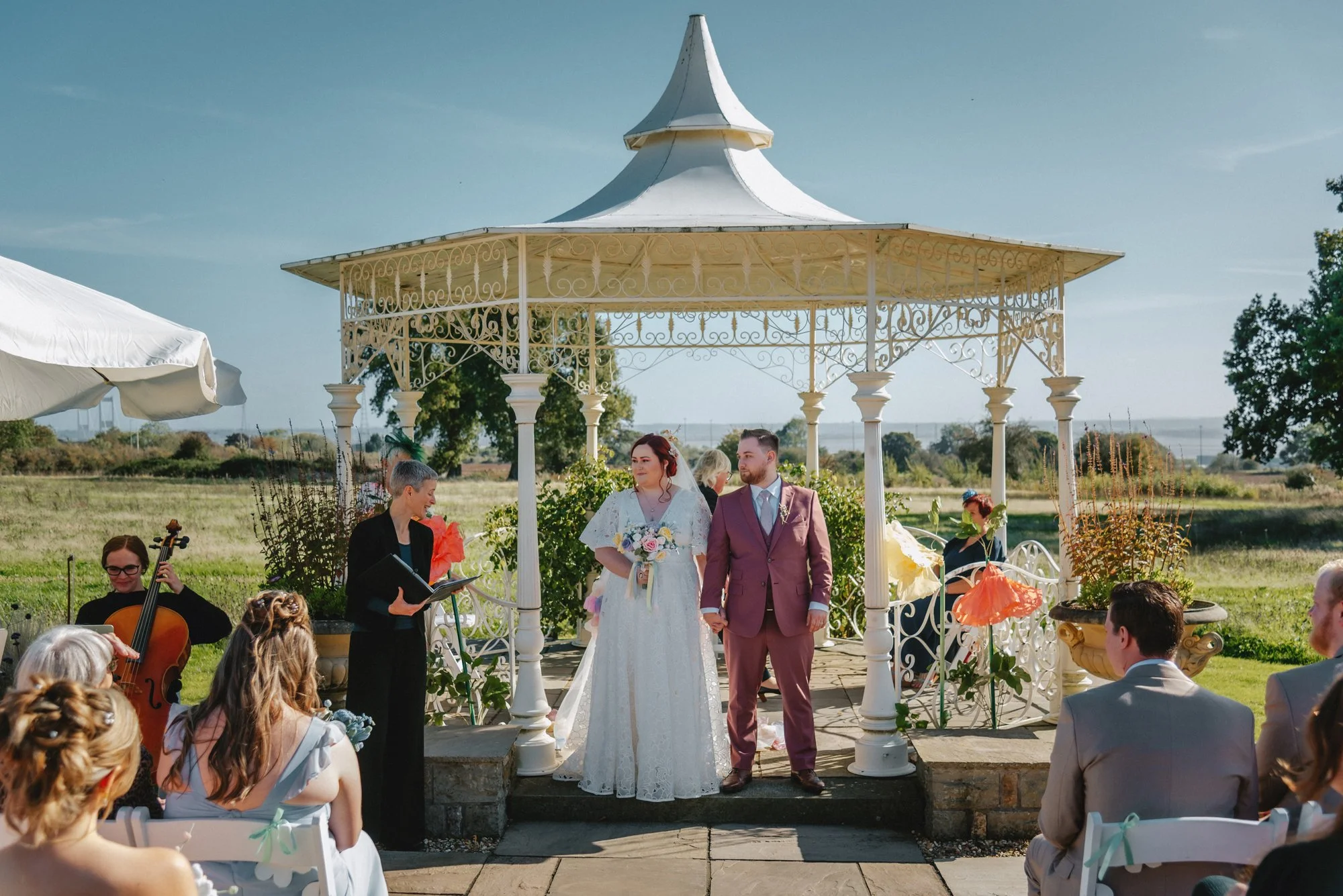 A bride and groom standing hand in hand during their outdoor wedding ceremony at a white ornate gazebo, surrounded by guests and musicians, on a sunny day with a rural landscape in the background.