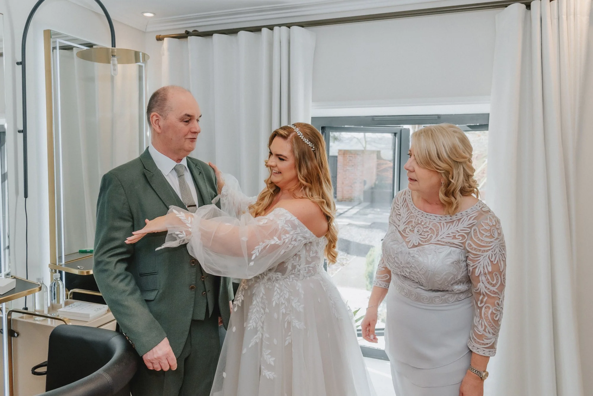 A bride in a wedding dress hugging a man in a suit while a woman in a lace dress looks on indoors with white curtains and a window.