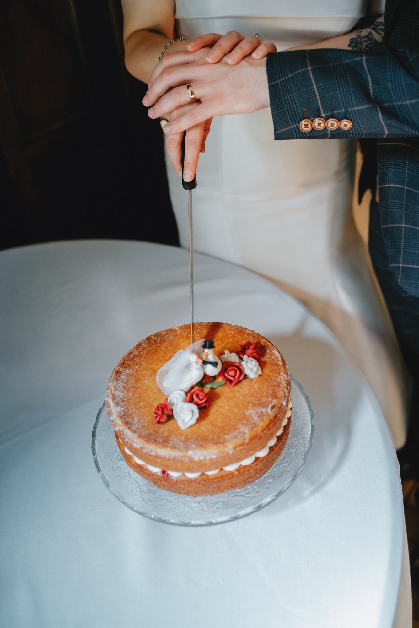 Couple cutting a wedding cake decorated with red and white flowers, with a cake knife, on a white tablecloth.