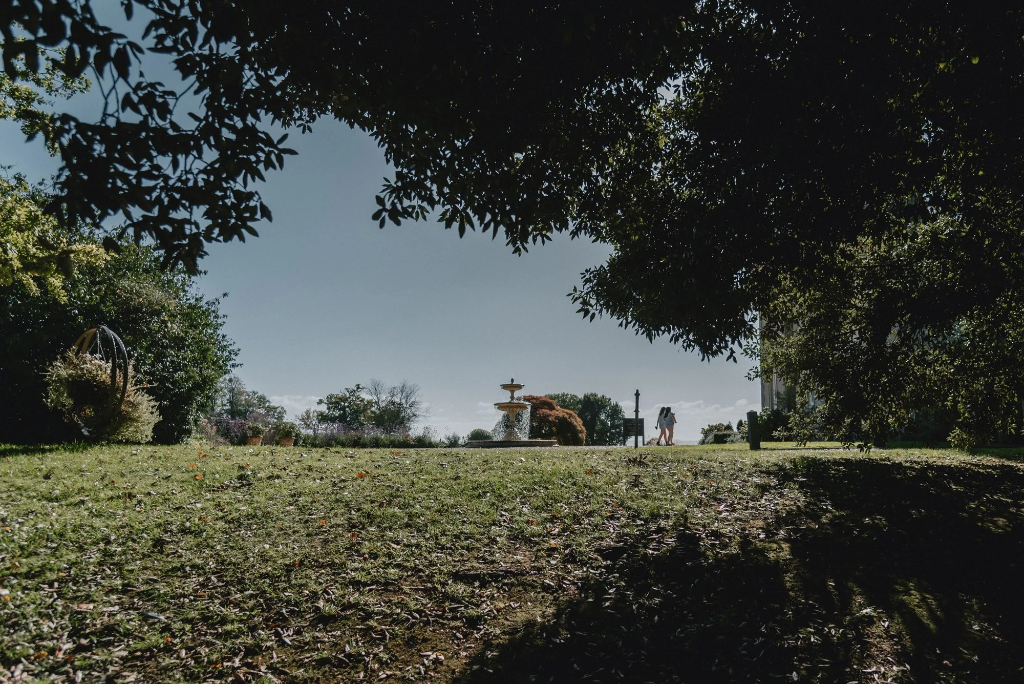 Two people walking along a grassy area near a fountain with trees and shrubs in the background.