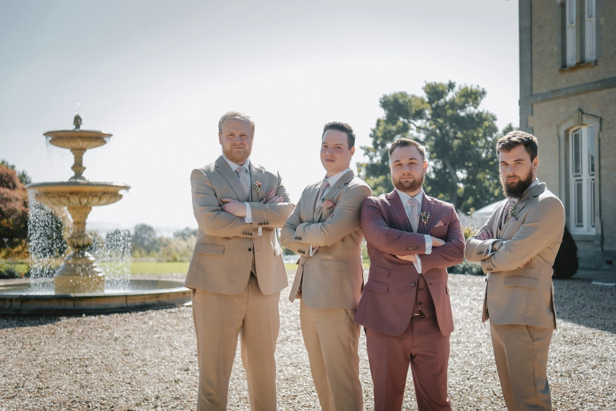 Four men in suits standing outdoors near a fountain and a large building, with greenery and trees in the background.