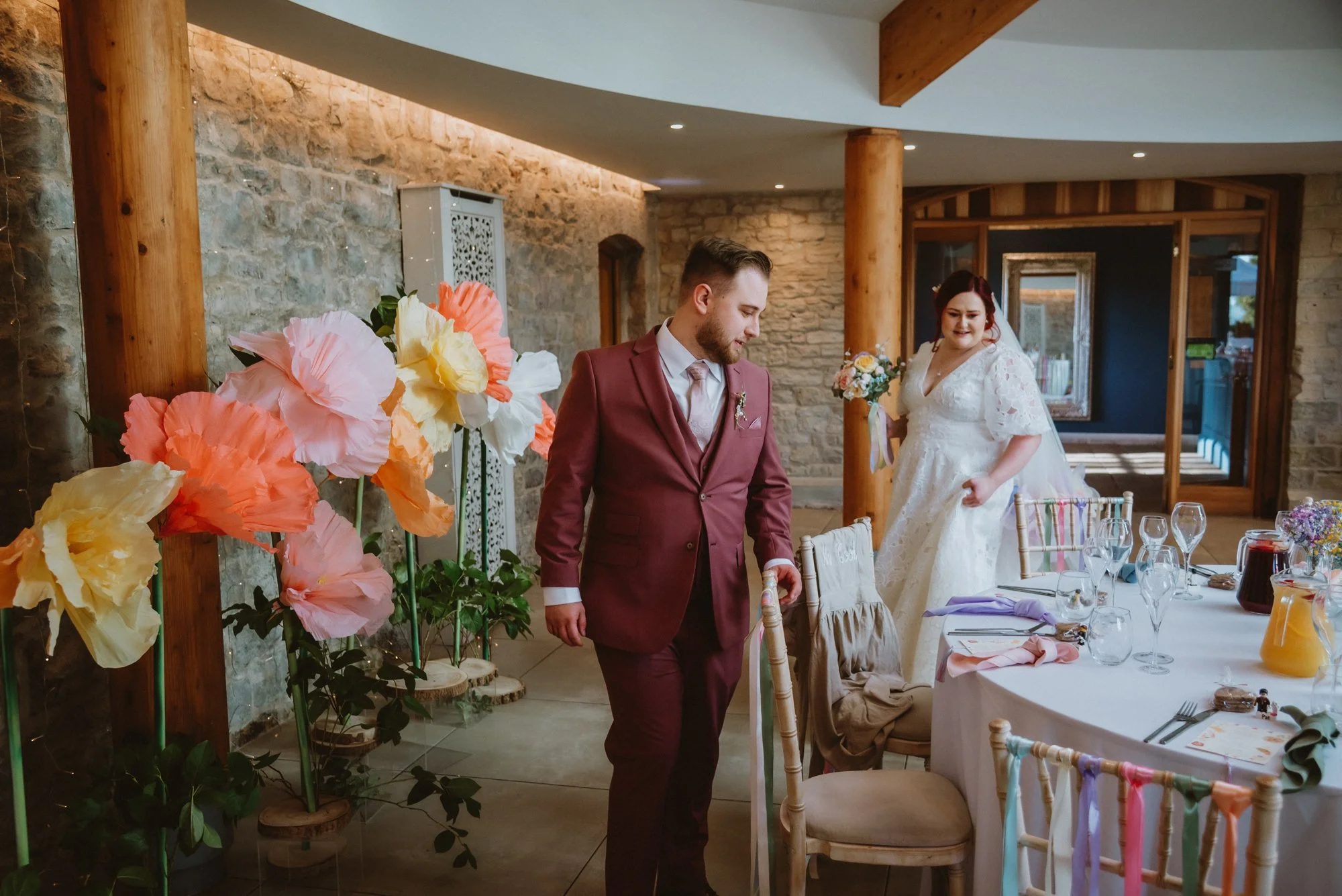 A wedding reception scene with a bride and groom, decorated with large paper flowers in pink, orange, and yellow, in a rustic-style venue with stone walls and wooden beams. The groom is dressed in a burgundy suit, while the bride wears a white lace w