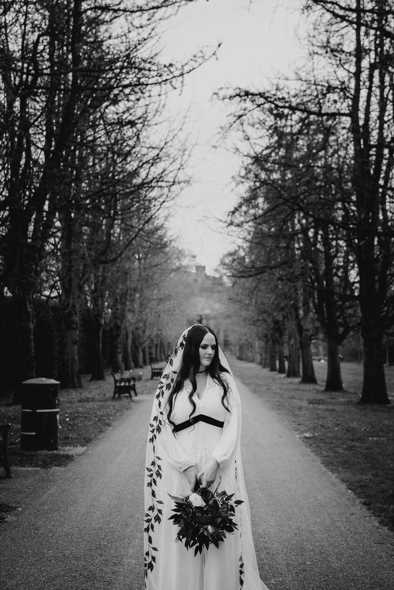 A woman in a wedding dress standing on a tree-lined path in a park, holding a bouquet of flowers.