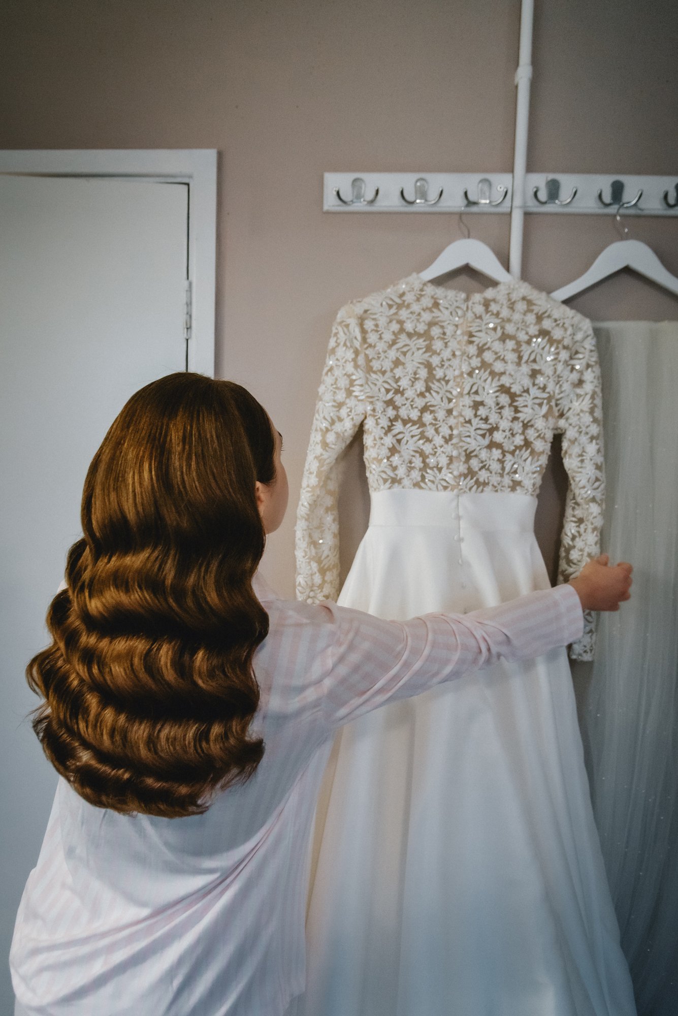A woman with long, wavy brown hair wearing a striped shirt is adjusting a white wedding dress that has a lace top with floral patterns and a satin skirt, hanging on a rack against a beige wall.