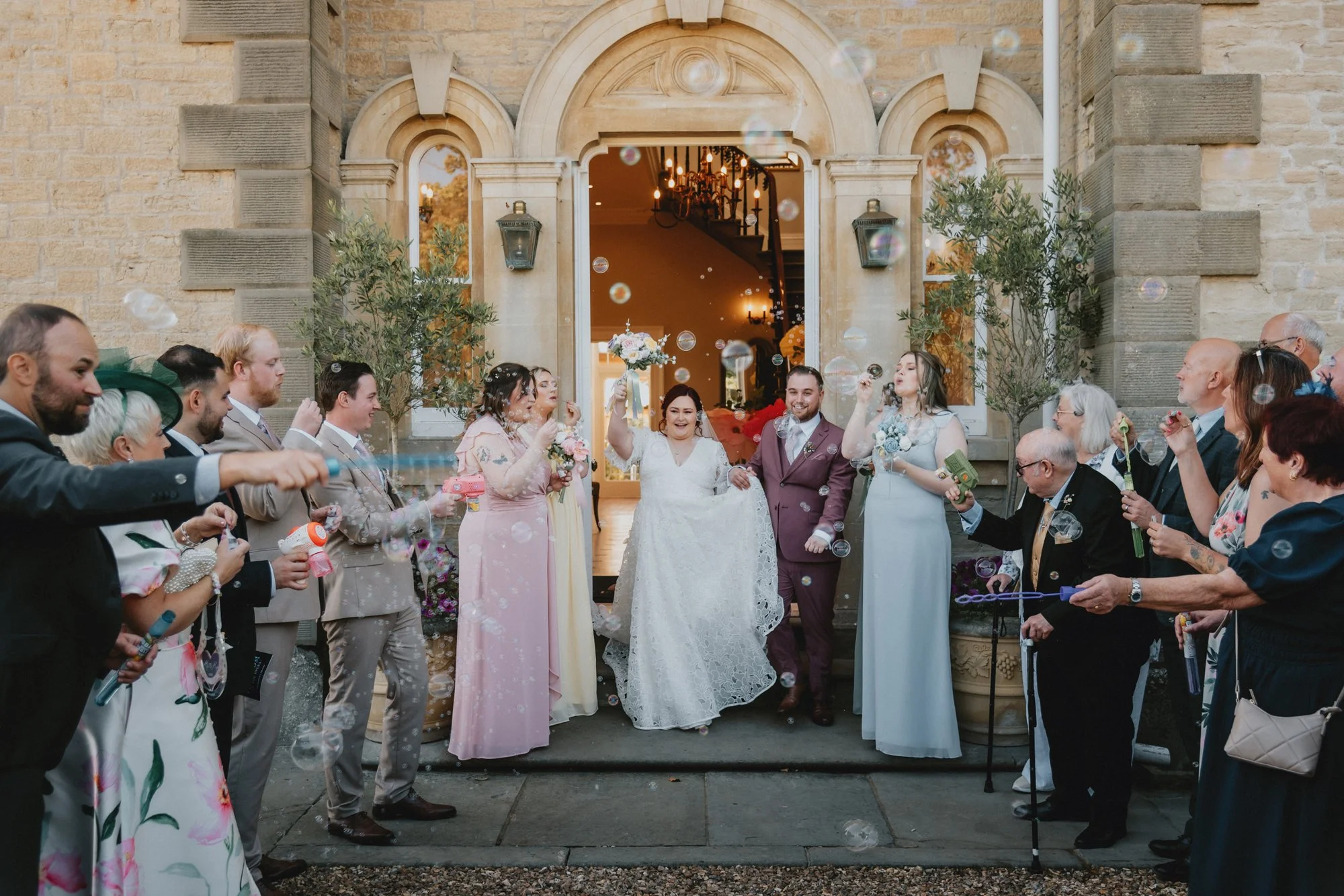 A wedding celebration outside a building with stone walls and an arched doorway, with the bride and groom surrounded by guests blowing bubbles.
