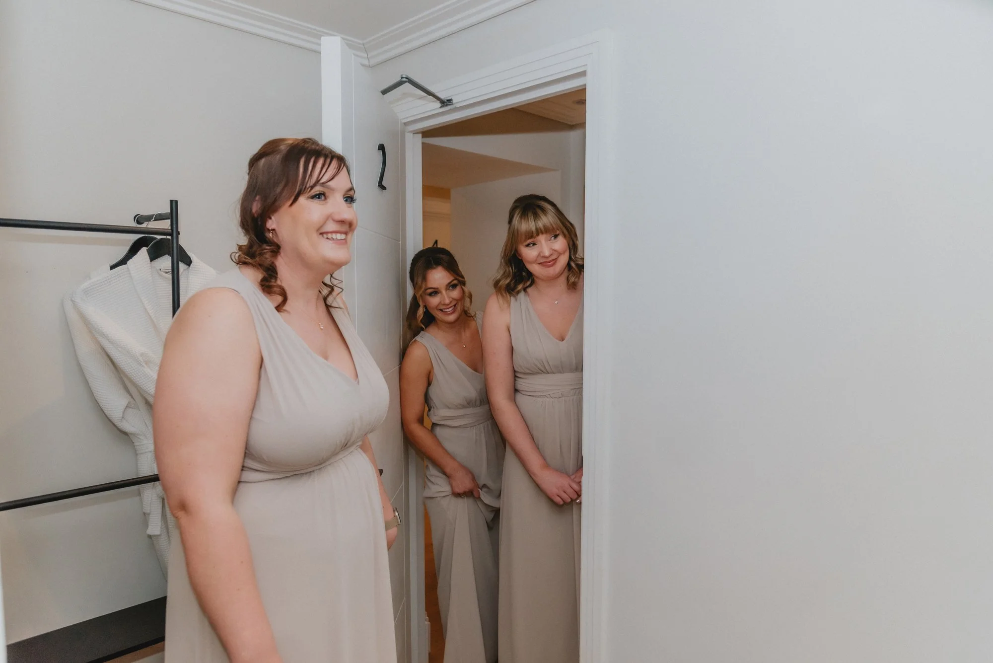 Three women in beige dresses smile and look out from a small room or closet at a wedding or formal event.