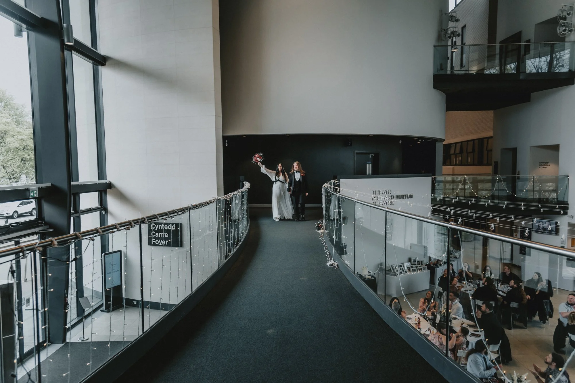 Two women, one in a wedding dress holding a bouquet, walking down an indoor corridor at a theater, with a balcony and dining area below. Decorations include fairy lights along the handrail.